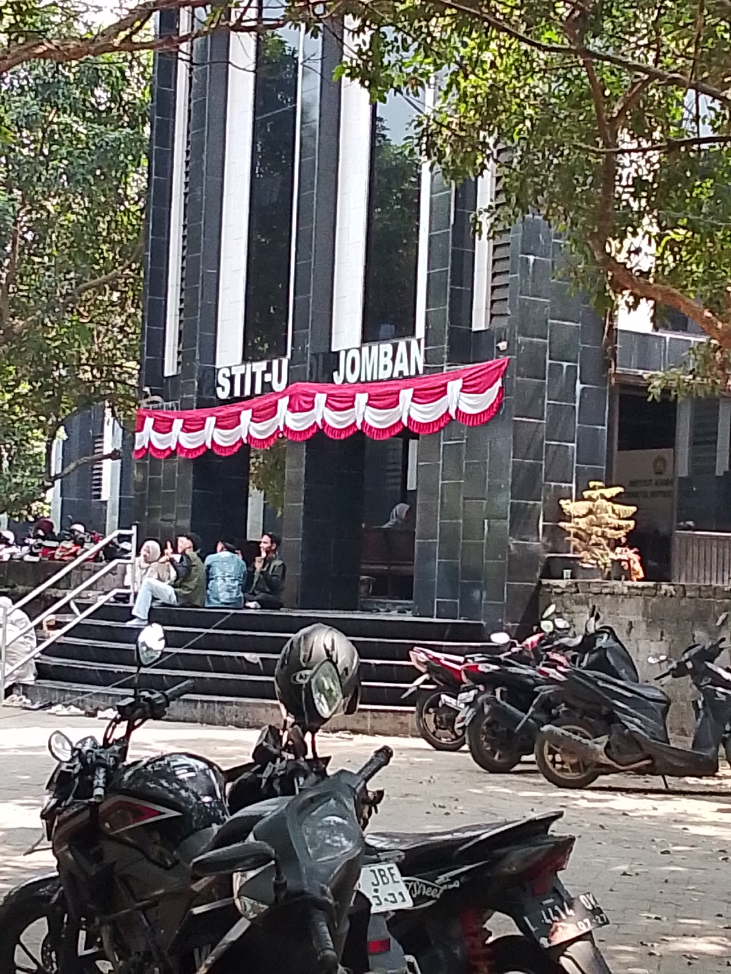 The image shows the exterior of a building with the text "STIT-U JOMBANG" visible. The building has a modern architectural style with dark gray stone cladding and vertical white accents. A red and white draped banner hangs across the front facade. In front of the building, a set of stairs leads up to an entrance. Several individuals are seated on the stairs, some appearing to be in conversation. The scene is outdoors, with trees providing shade. In the foreground, several motorcycles are parked. One motorcycle has a helmet placed on its handlebars. The overall impression is that of a daytime scene outside an educational institution.