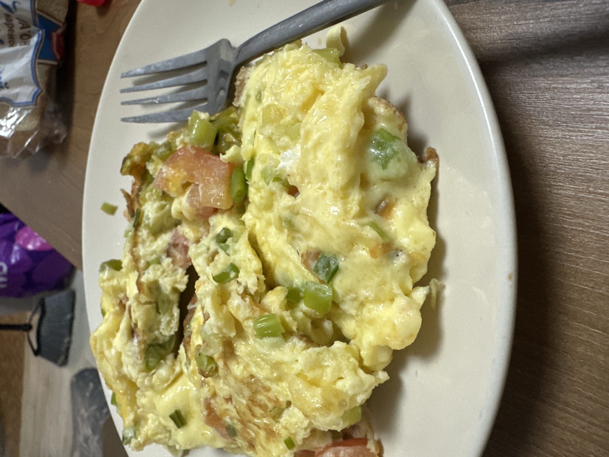 A plate of scrambled eggs with visible vegetables (likely green onions and tomatoes) is placed on a wooden surface. A fork rests atop the eggs. Part of a bag is visible on the left side of the frame. The specific location is not discernible from the image.