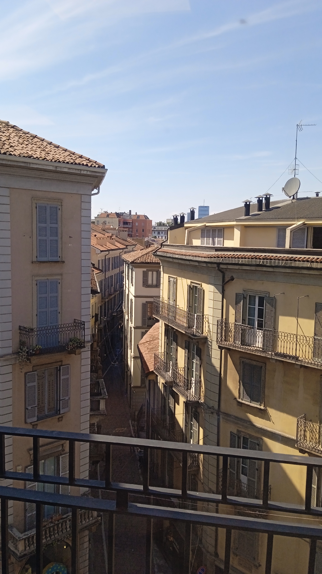 The image depicts a view from a balcony overlooking a narrow street lined with apartment buildings in Milan, Italy. The buildings are of various heights and architectural styles, with many featuring terracotta tiled roofs and shuttered windows. Balconies with ornate iron railings are visible on several of the buildings. The sky is clear and blue with a few wispy clouds. The sun is casting shadows on the buildings, indicating it is daytime. In the distance, taller, modern buildings can be seen, suggesting an urban environment. There are no people visible in the scene. The overall impression is of a typical residential street in an Italian city.