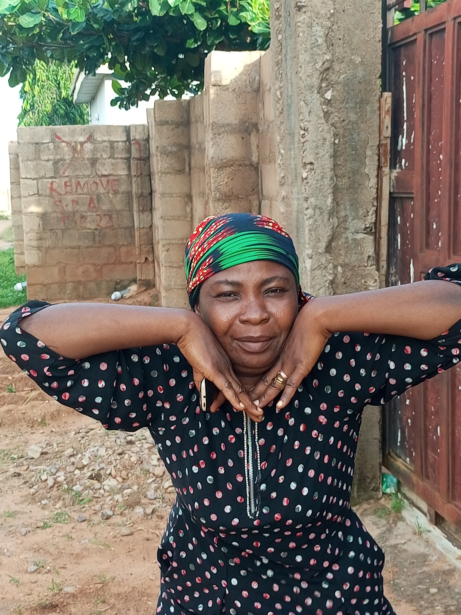A woman with dark skin and a headscarf covering her hair and neck is posing for a photo. The headscarf has a geometric pattern of red, green, and black. She is wearing a black dress with a pattern of colorful polka dots. Her hands are resting on her shoulders, and she is looking at the camera. The background suggests an outdoor setting in Tempure, Nigeria, with a rough concrete wall and a reddish-brown metal gate. There are also some brick structures and foliage visible. The ground is covered with dirt and gravel. The overall impression is of a candid portrait taken in a casual, possibly residential or semi-industrial, area.