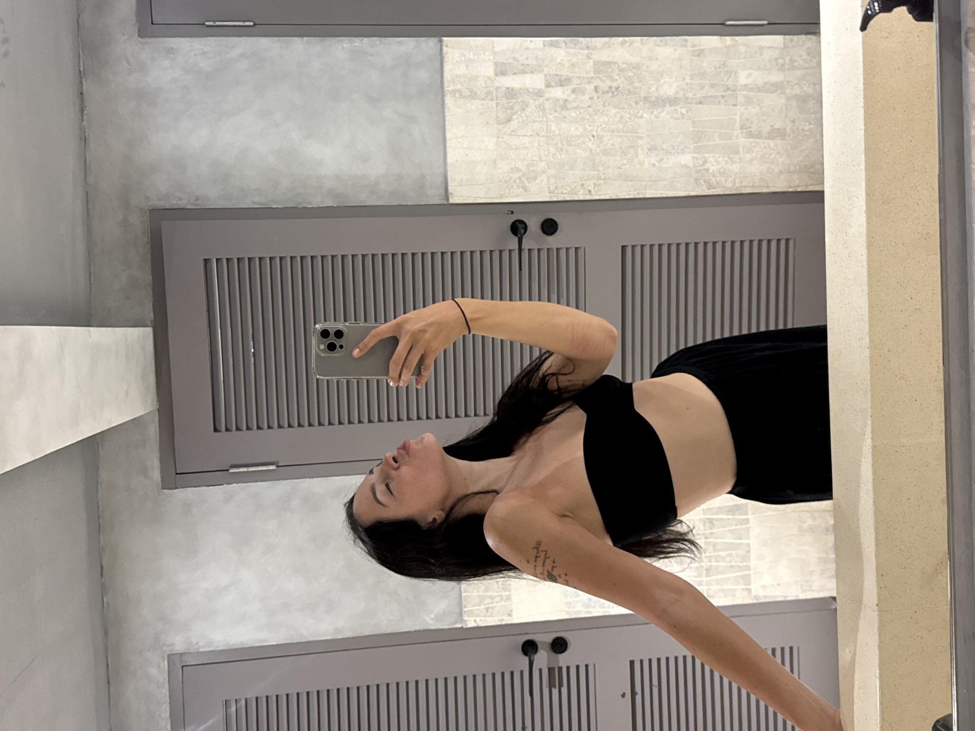 A woman with long, dark hair is taking a selfie in a mirror. She is wearing a black top and black pants. Her hair is partially obscuring her face as she looks at the camera. A phone is held in her right hand, and her left arm is extended. A tattoo is visible on her left arm. The background appears to be a bathroom or dressing room with grey walls and cabinets with vertical slatted doors. The lighting suggests it might be daytime, and the overall impression is casual. The location is North Kuta, Indonesia.