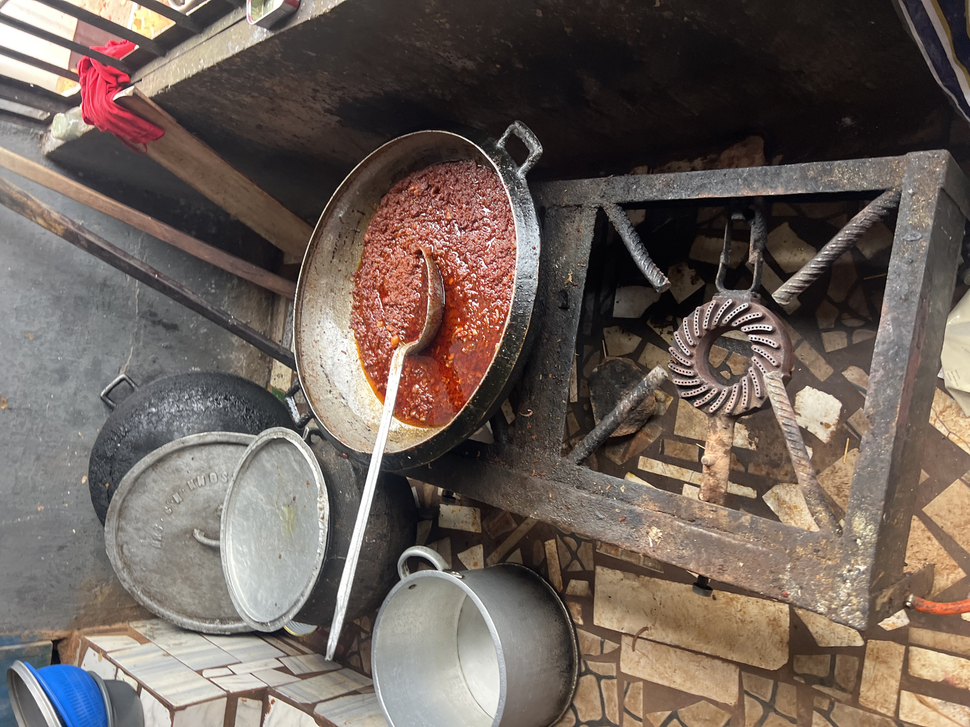 The image depicts an outdoor cooking scene in Nkpor, Nigeria. A large black pot with a lid is placed on a rustic, metal stove. To the left of the stove, a metal pot with a handle sits on the ground, and a blue and gray colander is stacked on top of a tiled surface. A wok-like pan filled with a reddish-brown stew is placed directly on the stove. A metal ladle is submerged in the stew, and a portion of a red cloth is visible in the upper right corner. The ground is tiled with irregular brown and cream-colored stones. The background consists of a textured wall and a corrugated metal structure. The overall impression is one of a functional, albeit somewhat weathered, outdoor kitchen.