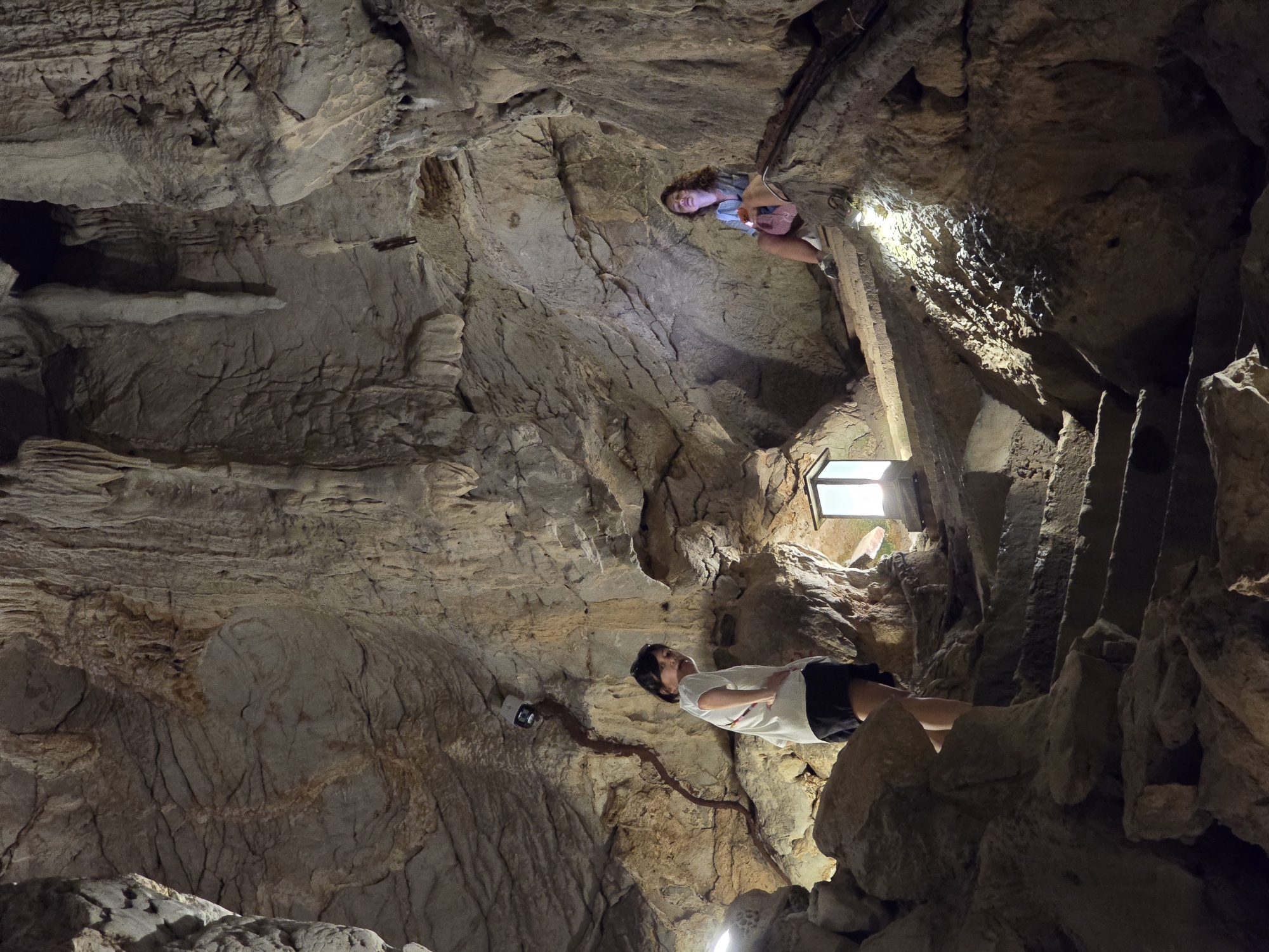 The image shows two young women inside a cave with stone formations. A stone staircase ascends from the bottom of the frame towards a landing where a lantern is placed. The woman on the left is standing on the staircase, wearing a white t-shirt and black shorts, and looking towards the camera. The woman on the right is sitting on the landing, wearing a denim jacket and shorts, and holding a phone. The cave walls are textured with stalactites and stalagmites, and artificial lighting illuminates the scene. The location is identified as Unknown, Unknown.