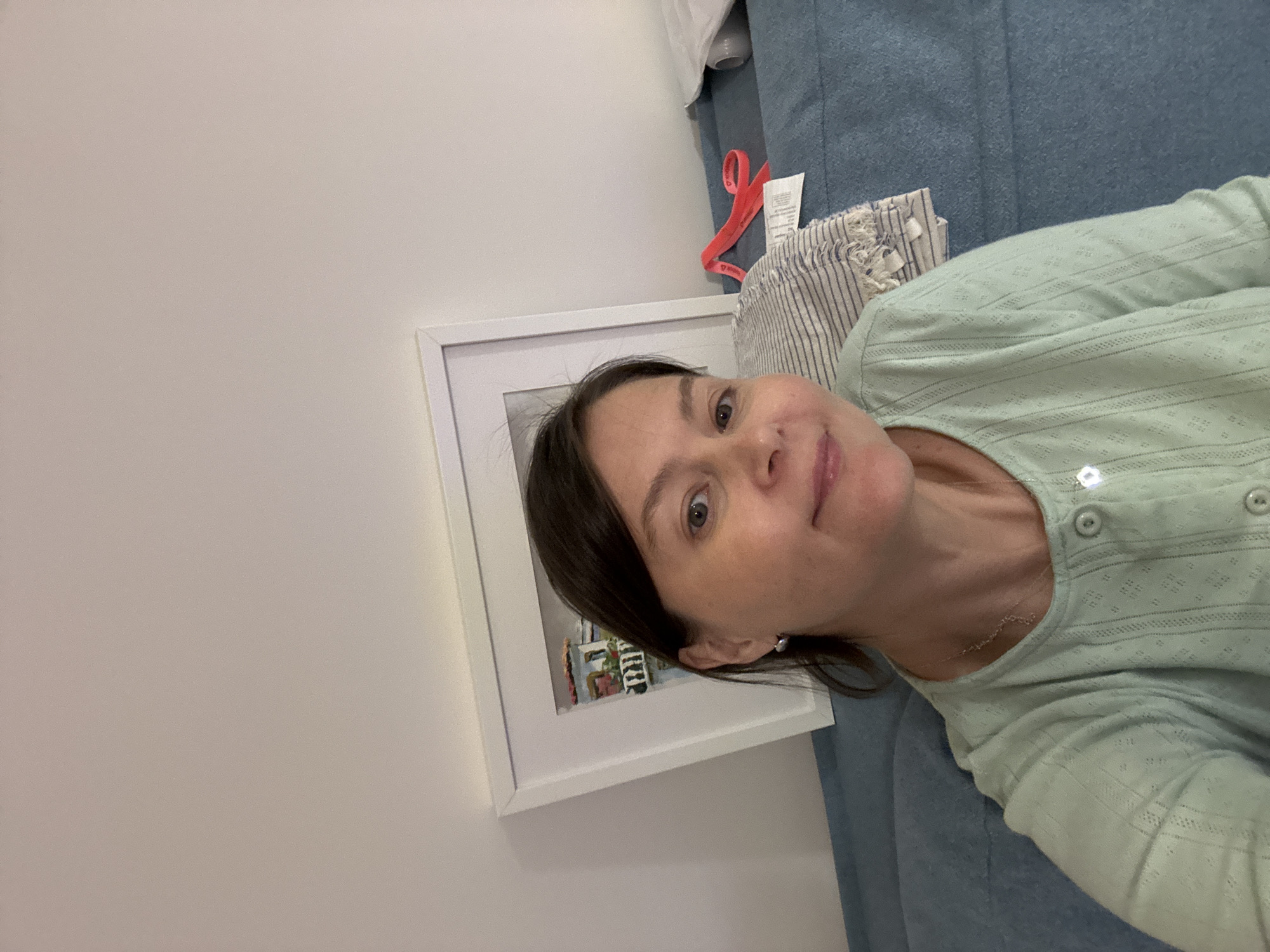 A medium close-up selfie captures a woman with dark brown hair and light eyes, looking directly at the camera with a soft, friendly smile. She appears to be reclining or lying down, with her head resting on a textured, light blue-grey fabric, likely a couch or bed. She is wearing a light mint-green, long-sleeved top featuring a subtle, patterned fabric with small eyelets and horizontal stripes, and has light-colored buttons down the front placket. A delicate silver-toned necklace and small pearl-like earrings are visible.
The background shows a plain white wall on the left, with a white-framed picture partially visible. The small artwork inside the frame depicts abstract, colorful shapes that could represent buildings or a street scene. To the right and behind the woman, more of the light blue-grey fabric is seen, along with a piece of white and blue striped fabric, possibly a cushion or blanket. A bright red ribbon or strap is tucked into the fabric in the upper right corner, along with a small white product label, the text of which is unreadable. The setting suggests an indoor environment, likely a home, in Izhevsk, Russia. The lighting is soft and even.
