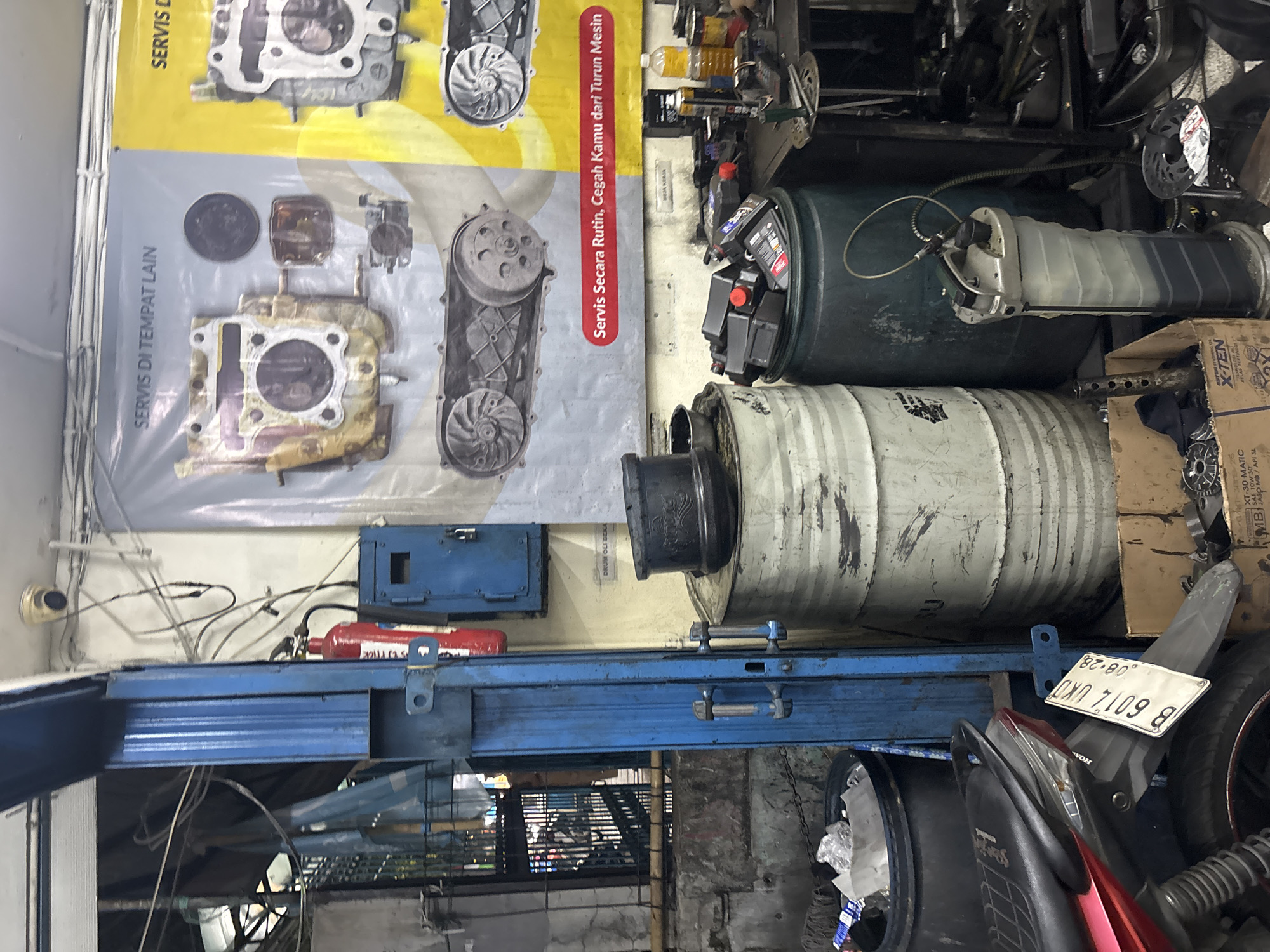 This image depicts the interior of a motorcycle repair shop, possibly located in North Jakarta, Indonesia. The scene is cluttered and industrial, with various parts, tools, and equipment visible. In the foreground, a red motorcycle is partially visible, with its license plate displaying "B 6017 UKU" and "08-28".
Dominating the mid-ground are two large, weathered drums. The one on the left is white and appears to be a used oil drum, with a black rim on top. The drum on the right is dark green and is filled with numerous black oil bottles. To the right of these drums, a gray fluid extractor is positioned, with a hose and collection tank.
In the background, a large banner hangs on the wall featuring diagrams of motorcycle engine parts and text that translates to "Regular Service, Prevent Engine Failure". Another banner to the right is partially visible. Adjacent to the blue support pillar, a red fire extinguisher is mounted on the wall next to a blue metal cabinet with a padlock.
Other objects in the scene include various tools, spare parts scattered on the floor, and a trash bin overflowing with crumpled plastic. The overall impression is one of a busy, working environment with a focus on motorcycle maintenance.