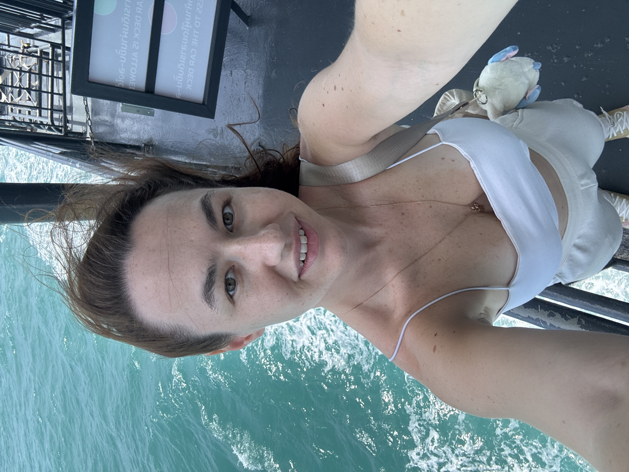 A woman with brown hair and green eyes, wearing a white tank top and light-colored pants, takes a selfie on a ferry with turquoise water visible to her left. She is smiling and her hair is blowing in the wind. A small, white plush toy with blue accents is clipped to her bag. Signs with Thai script are visible in the background. The scene appears to be on a ferry traveling in Thailand.