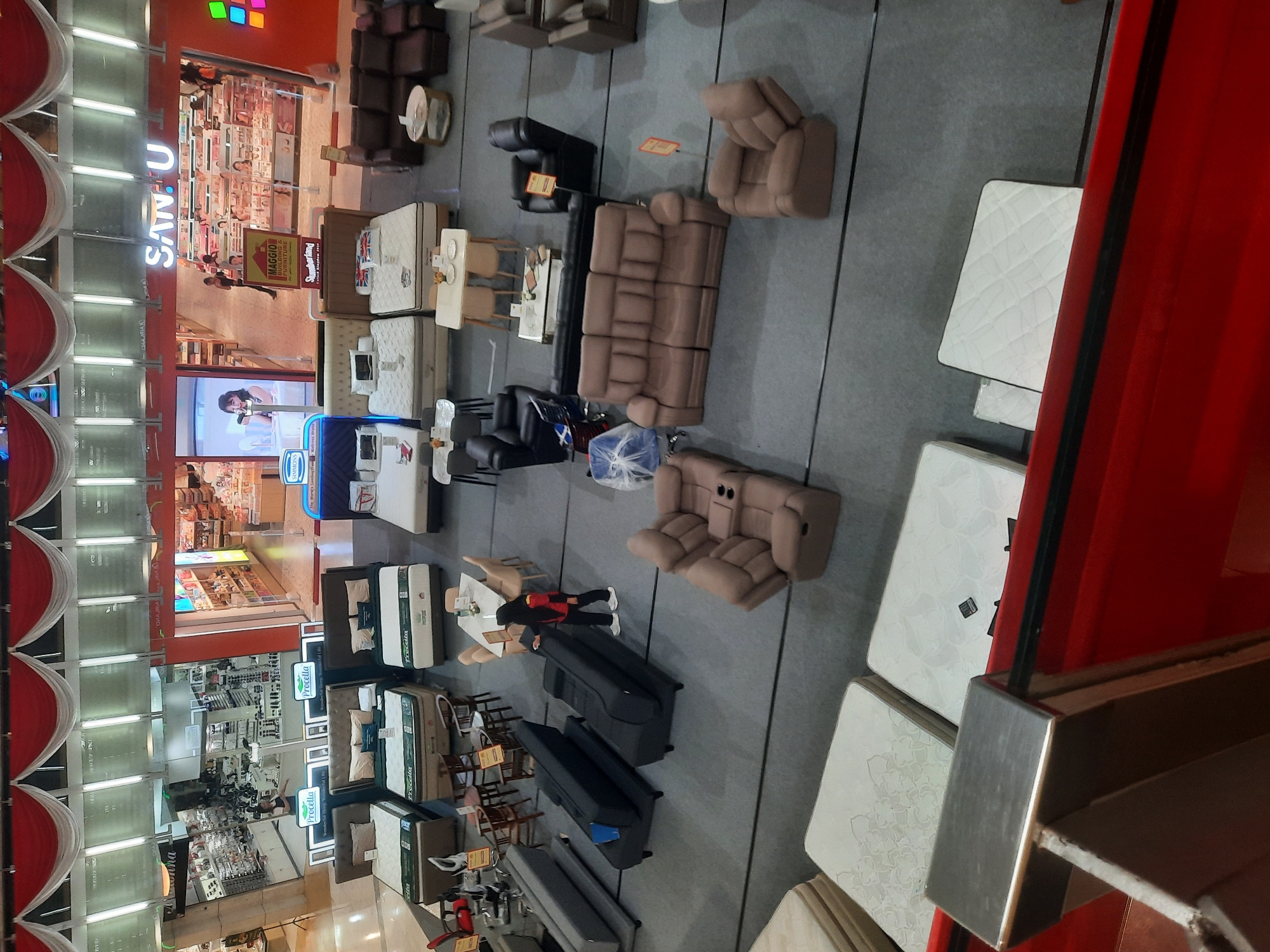 This image depicts the interior of a furniture store, likely a showroom, located in the city of North Jakarta, Indonesia. The store features a wide array of furniture, including numerous beds and mattresses displayed on the floor, and various seating arrangements such as sofas and armchairs. The layout suggests a retail environment, with products arranged for customer viewing and potential purchase.
In the background, there are store entrances leading to other sections, possibly for different brands or product categories. One prominent sign reads "SAN-U." Several individuals are visible in the scene, some appearing to be customers browsing, while others may be store employees. One person in the foreground is interacting with a black sofa, possibly arranging it or cleaning. The environment is well-lit, consistent with a commercial space. The overall impression is that of a busy retail establishment.