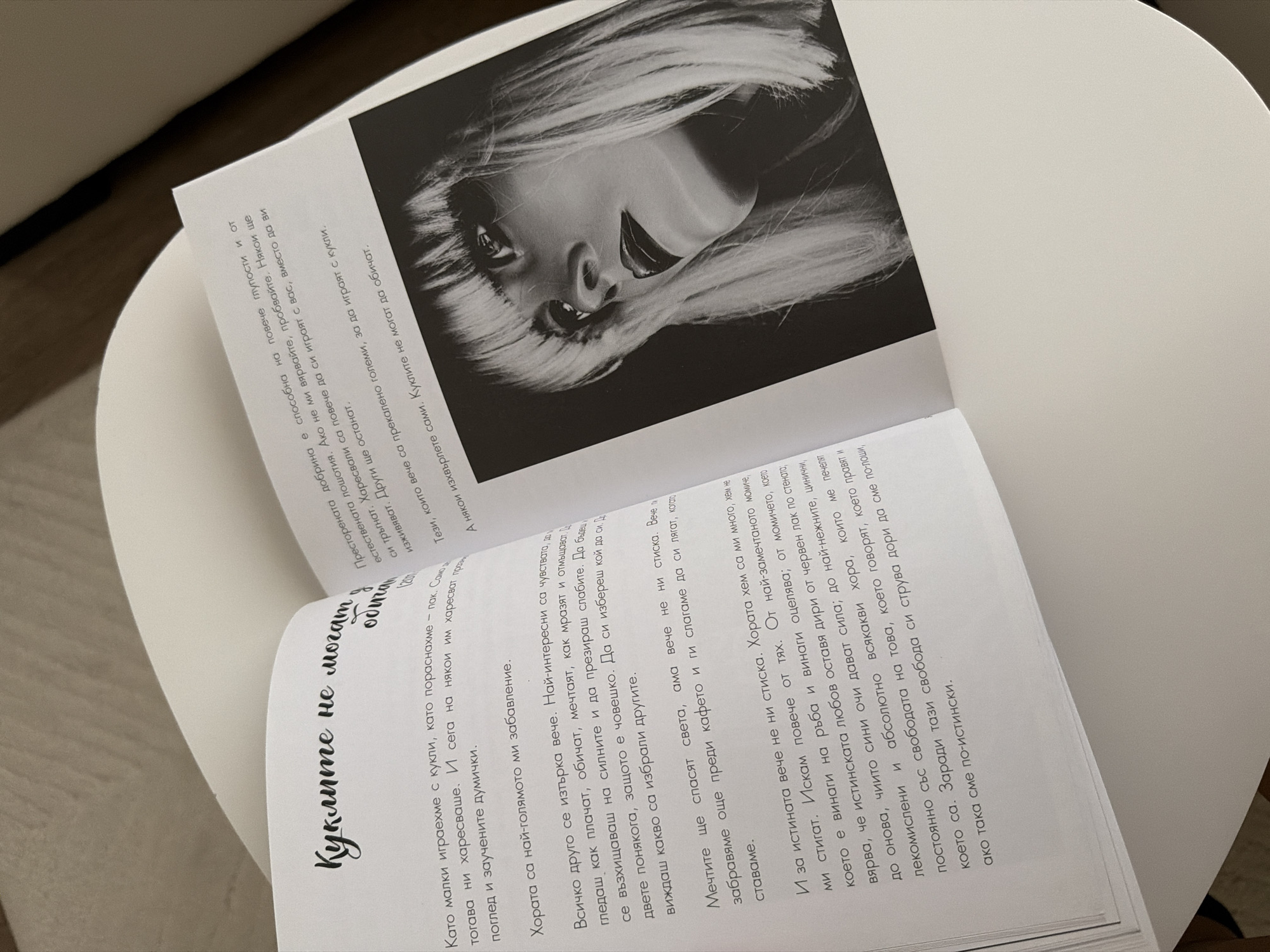 The image shows an open book with text and a black and white photograph. The text is in Bulgarian and appears to be a personal reflection or essay. The photograph is a close-up portrait of a woman with blonde hair and dark lipstick, looking to the side.
The book is lying on a flat surface, possibly a table, and the lighting suggests it is indoors. The book's pages are slightly curved, indicating it is newly opened or of a flexible binding. The photograph occupies the right half of the open book, partially obscuring the text on that side.
The text on the left page discusses childhood memories of playing with dolls and how relationships change with age. The text on the right page, partially visible, discusses how people are capable of love and forgiveness, and how some relationships are discarded.
There is no information within the image that would indicate its location in Pazardzhik, Bulgaria.