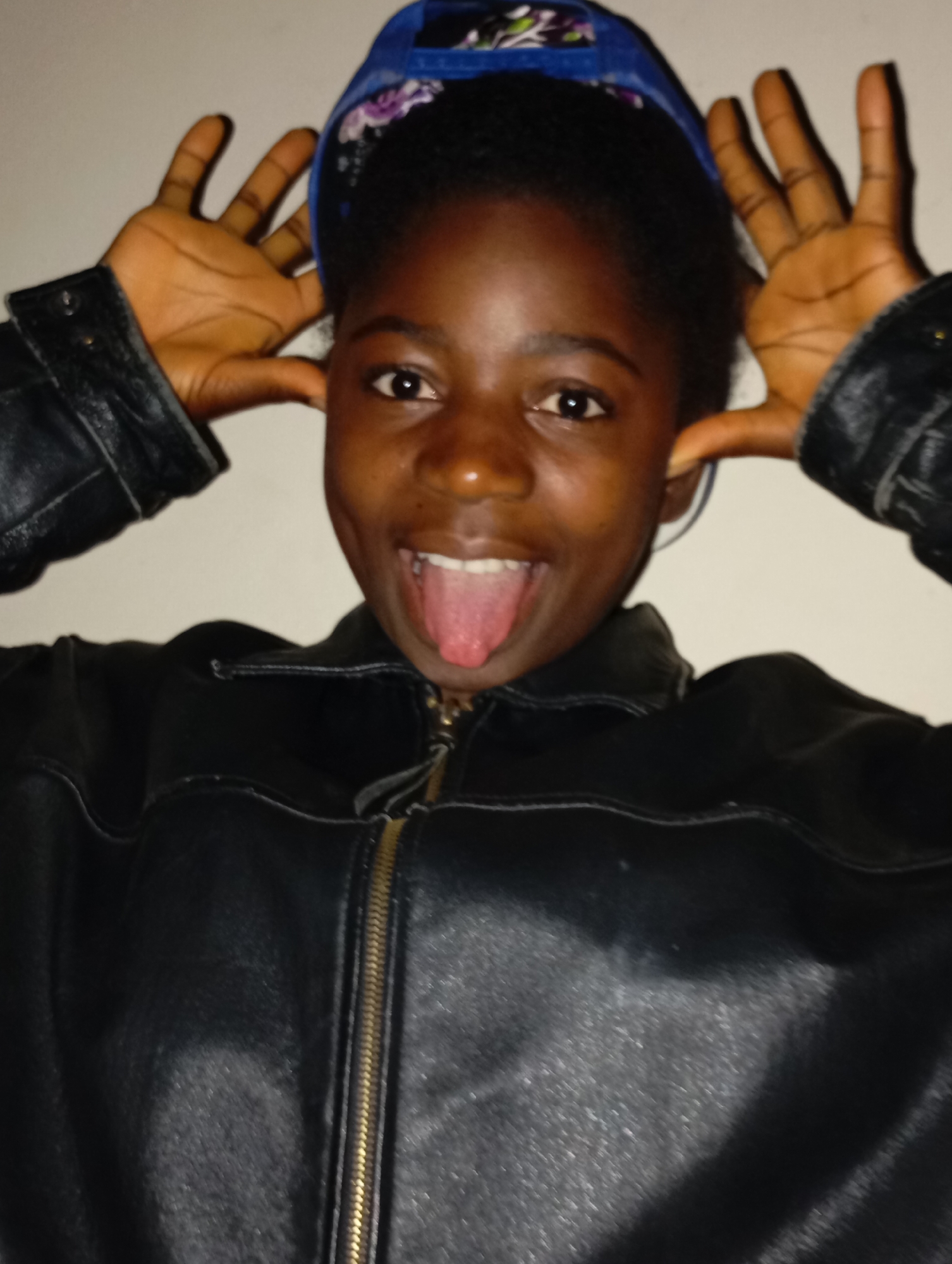 A young person with dark skin and a black leather jacket is pulling their ears with their hands and sticking out their tongue. They are wearing a blue baseball cap with a floral pattern on the brim. The background is a plain, light-colored wall. The location is Unknown, Unknown.