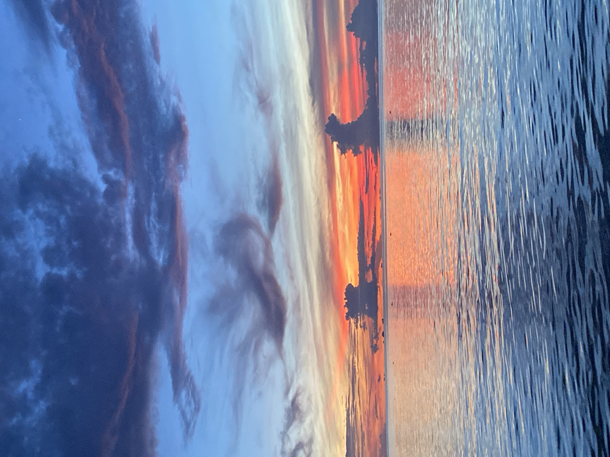 A sunset over the ocean in Koh Phangan, Thailand. The sky is a blend of deep blues, purples, and vibrant oranges and reds, with dramatic, silhouetted clouds. The water reflects the colors of the sky, creating a shimmering surface. The horizon line is distinct, with several small islands or landmasses visible in the distance. The overall scene is serene and picturesque.