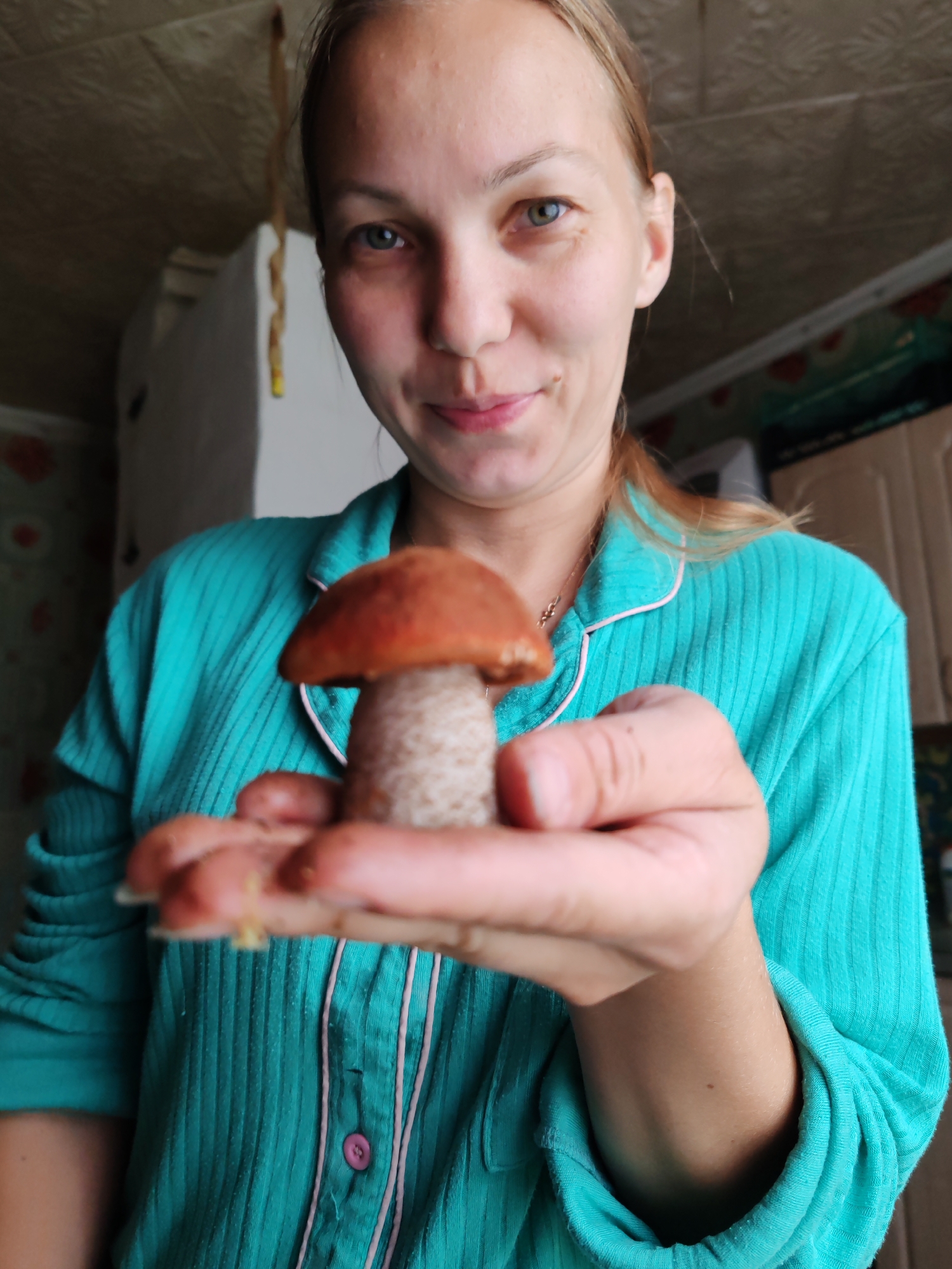 A young woman with blonde hair and light-colored eyes is holding a single small mushroom, specifically an orange-cap boletus, in her outstretched palm. She is wearing a turquoise pajama top with pink trim. The background appears to be an indoor domestic setting, possibly a kitchen, with cabinets and a ceiling visible. The image is taken from a close-up perspective, focusing on the woman and the mushroom. There is no visible indication of the location being in the city of Чухлэм, Russia.