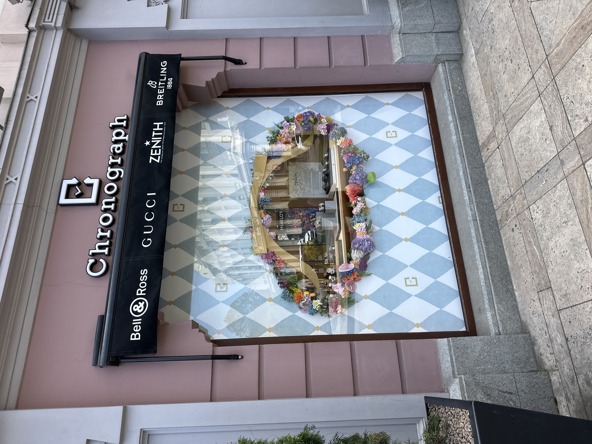 A clear daytime shot captures the storefront of a luxury watch and jewelry store named "Chronograph" on a city street in Tbilisi, Georgia. The building's facade is painted a light pink or dusty rose color, with white architectural details like cornices and pilasters.
The prominent feature is a large display window showcasing various luxury watch brands. Inside the window, a large, ornate frame, possibly gilded, is elaborately decorated with a vibrant array of artificial flowers in various colors, including purple, pink, orange, yellow, and white, forming an irregular, almost oval-shaped floral wreath. The background behind the floral display is a light blue and white argyle or diamond pattern with small golden square-like symbols in the white diamonds. Reflections in the window show parts of the street and possibly the store's interior, including faint text like "HUBLOT" and "ZENITH."
Above the window, a black retractable awning or banner displays several luxury brand names vertically: "GUCCI", "Bell & Ross", "ZENITH" (with "THE FUTURE OF SWISS WATCHMAKING SINCE 1865"), and "BREITLING 1884." To the left, large white three-dimensional letters spell "Chronograph," accompanied by a distinct logo resembling a stylized 'B' or stopwatch.
The ground in front of the store consists of grey, textured stone paving, with a dark planter box containing small green plants visible in the lower right. The scene suggests a sophisticated commercial area, likely bustling during the day, with no people visible in the immediate shot.