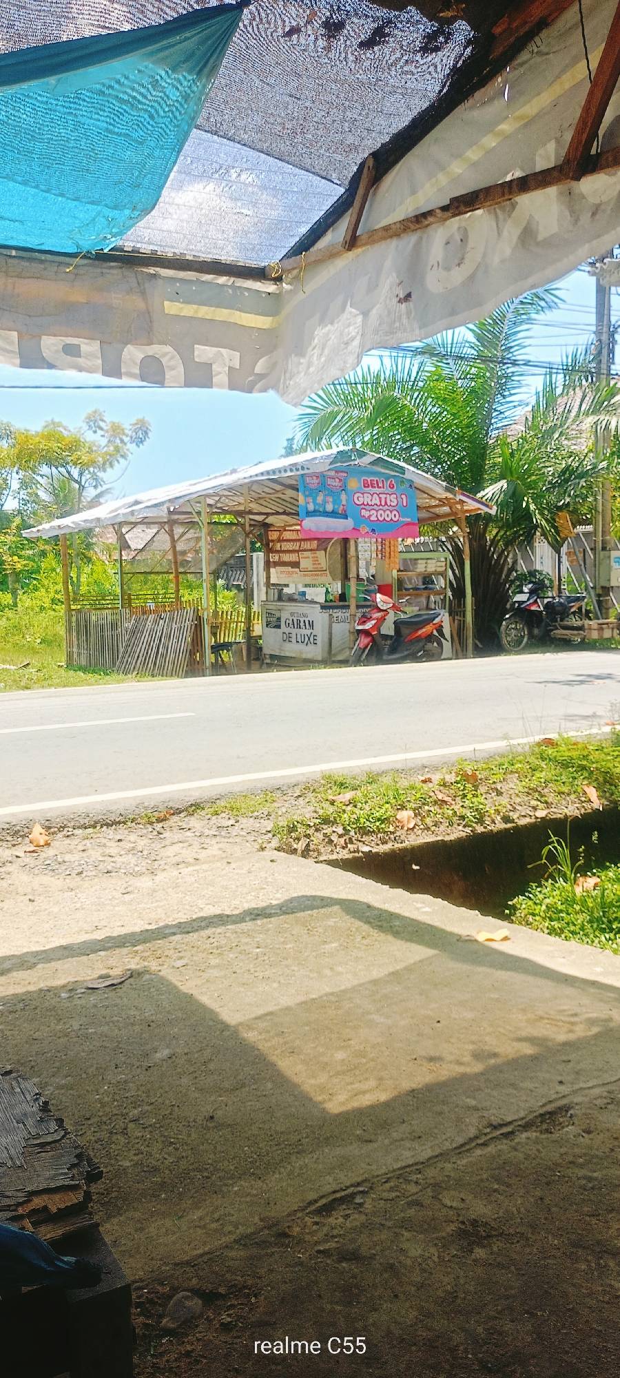 The image depicts a roadside stall in Lhokseumawe, Indonesia, under a makeshift canopy. The stall features a wooden structure with a corrugated metal roof, covered with a blue tarp and what appears to be netting. A banner hangs from the front of the stall, advertising a promotion: "BELI 6 GRATIS 1 RP 2000." Below this, another sign reads "GEDUNG GARAM DE LUXE." A red and white scooter is parked to the right of the stall.
Behind the stall, there is dense green vegetation and some bamboo fencing. A road runs horizontally across the mid-ground of the image. In the foreground, a concrete surface with shadows and a drainage ditch is visible. The overall scene suggests a warm, sunny day.