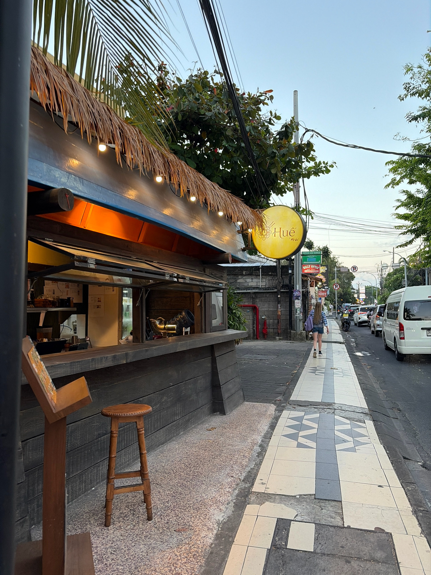 This is a street-level, eye-level medium shot of a sidewalk and a food stall in Kuta, Indonesia. The food stall has a thatched roof with string lights and a wooden counter. Behind the counter, a window is open, showing food preparation areas and equipment. A wooden bar stool sits in front of the counter. A sidewalk with a patterned tile design runs alongside the street, which is lined with vehicles. Trees and utility poles with wires are visible in the background. The sky is clear and light blue, indicating it is either early morning or late afternoon. A person with long, blonde hair is walking away from the camera on the sidewalk, wearing a blue and white checkered shirt and denim shorts. A yellow circular sign with the text "Hue" is hanging from a pole above the food stall.