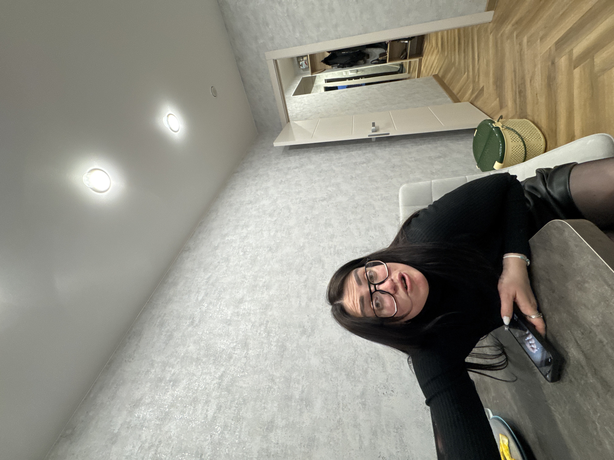 A woman is captured in a close-up, likely selfie-style, shot in a modern indoor setting. She is positioned on the right side of the frame, appearing to be lying on her side or leaning back on a light-colored sofa. She has long dark hair, wears dark-rimmed glasses, and is dressed in a black long-sleeved top and what appear to be dark leather shorts or a skirt, along with dark hosiery. Her expression is one of slight surprise or playfulness, with wide eyes and an open mouth. She holds a black smartphone in her right hand, though its screen content is indistinct. A light-colored beaded bracelet is visible on her left wrist, and she wears several rings on her fingers.
The room features a textured, light gray wallpaper on the wall behind her. The ceiling, visible on the upper left, is plain and light-colored, fitted with two brightly illuminated recessed spotlights and one unlit fixture further back. To the upper right, beyond the sofa, a wooden floor with a diagonal or herringbone pattern is visible. Below this, a light-colored built-in cabinet or console unit with a mirrored section reflects what appears to be an open wardrobe or shelving system filled with various items. A green and beige woven basket rests near the sofa. The overall lighting is artificial and bright, typical of an indoor environment. No discernible text is visible on any surfaces or the phone screen.