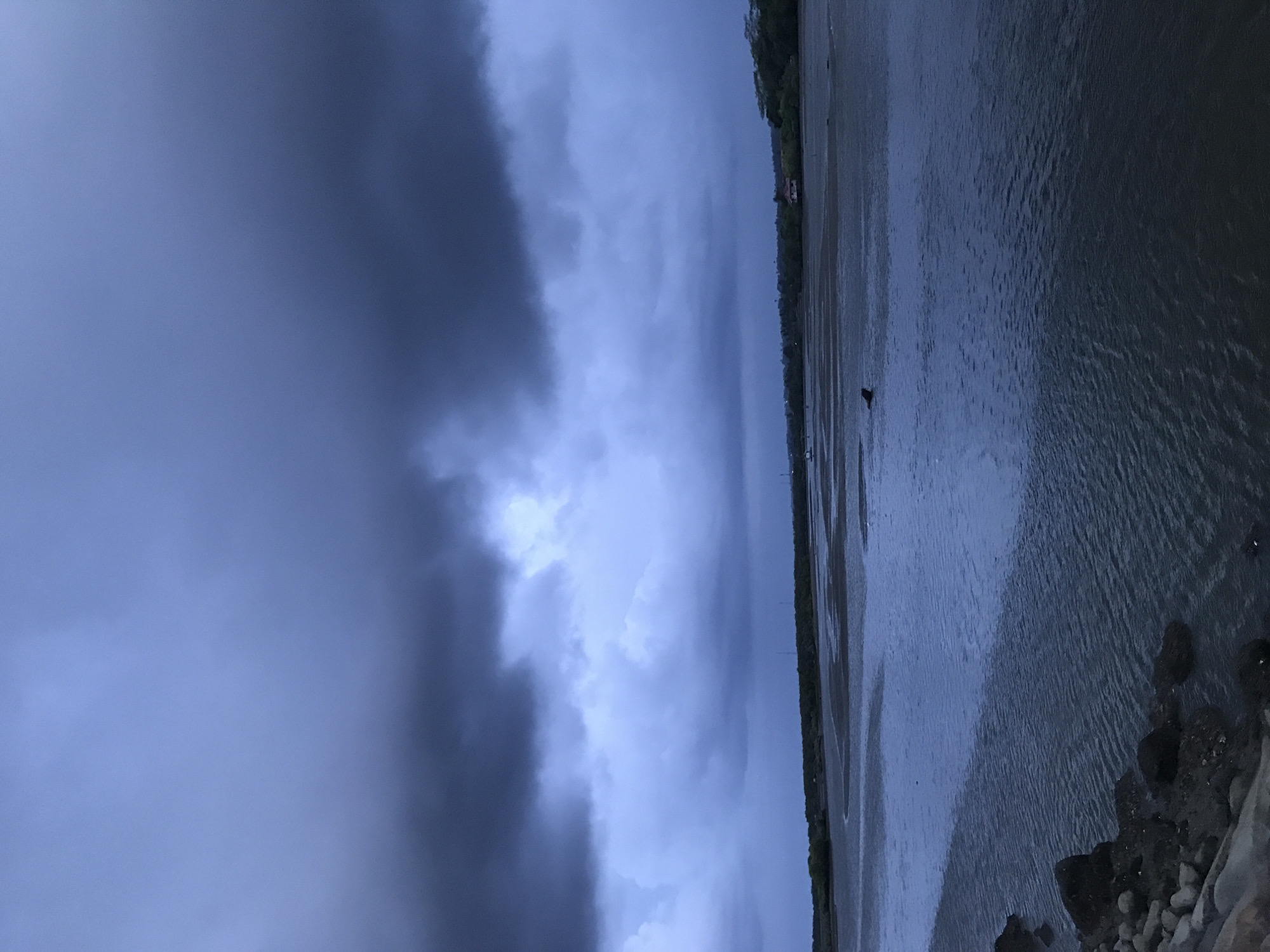 The image shows a landscape with a body of water, likely a river or bay, under a stormy, overcast sky. Dark, heavy clouds dominate the left portion of the image, suggesting impending or recent rainfall. A strip of land with vegetation, possibly a shoreline, runs along the horizon. The water's surface appears choppy, reflecting the dim light of the sky. In the foreground, there are rocks and stones.
The location is plausibly Lhokseumawe, Indonesia, based on the landscape and coastal characteristics of that region. There are no people visible in the image. The essential subjects present are water, sky, clouds, land, and rocks. No actions or interactions are directly observable, except the implied presence of weather. The environment is a coastal or riverside setting during a period of overcast or stormy weather.