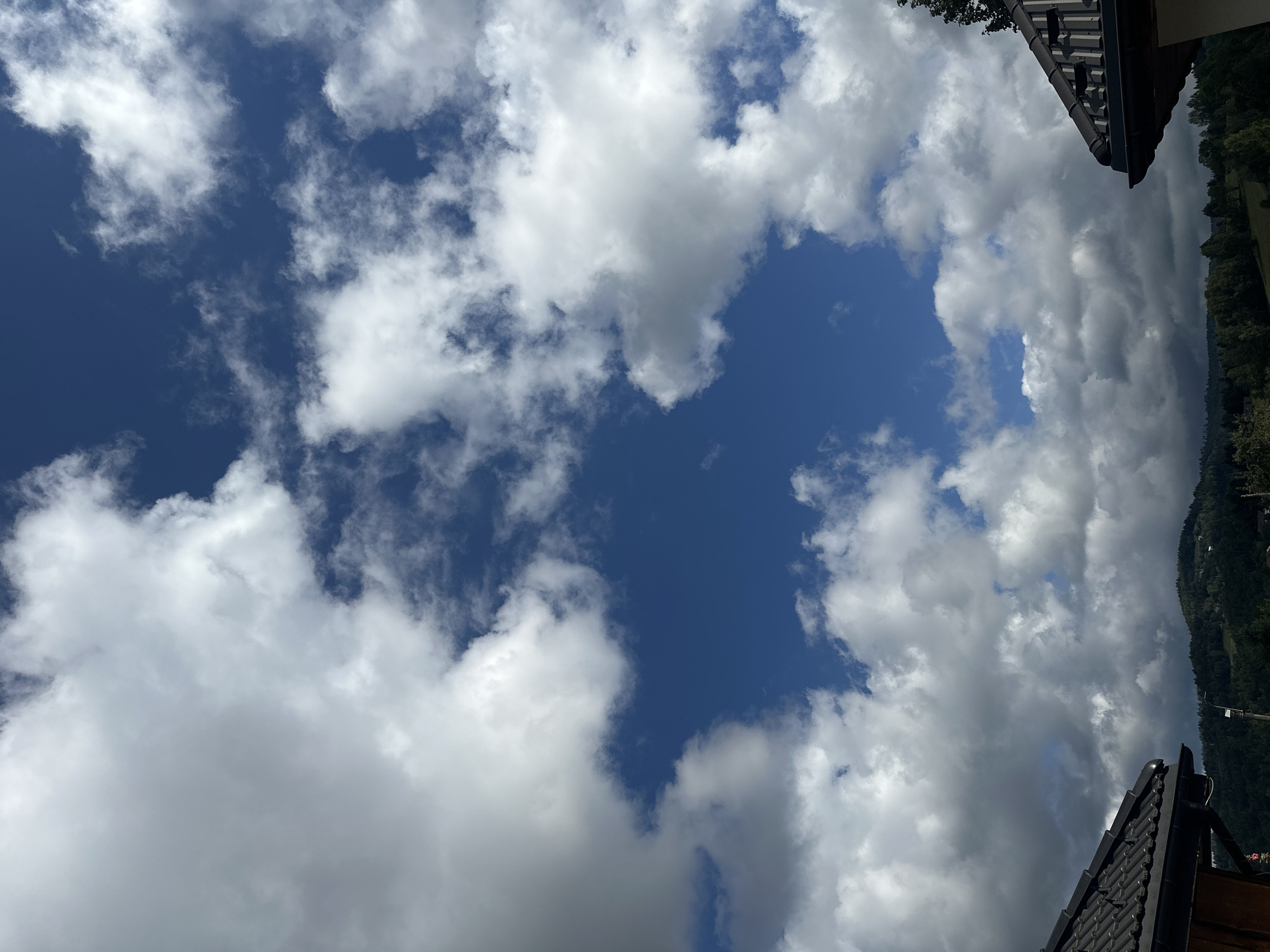 The image shows a cloudy sky over a hilly, forested landscape with two rooftops visible in the foreground. The sky is a deep blue with large, white cumulus clouds and scattered smaller, wispy clouds. The clouds are thick and appear to be casting shadows on the landscape below, suggesting a partly cloudy or overcast day.
The rooftops are dark, tiled structures, likely belonging to houses or other buildings. One rooftop is on the left side of the image, and the other is on the right. The landscape consists of rolling hills covered in dense green trees, indicating a natural, possibly rural or semi-rural environment. The overall impression is one of a natural landscape under a dynamic sky. The image does not contain any people or specific actions. The location is not explicitly identifiable as Wisła, Poland, based solely on the visual information in the image.