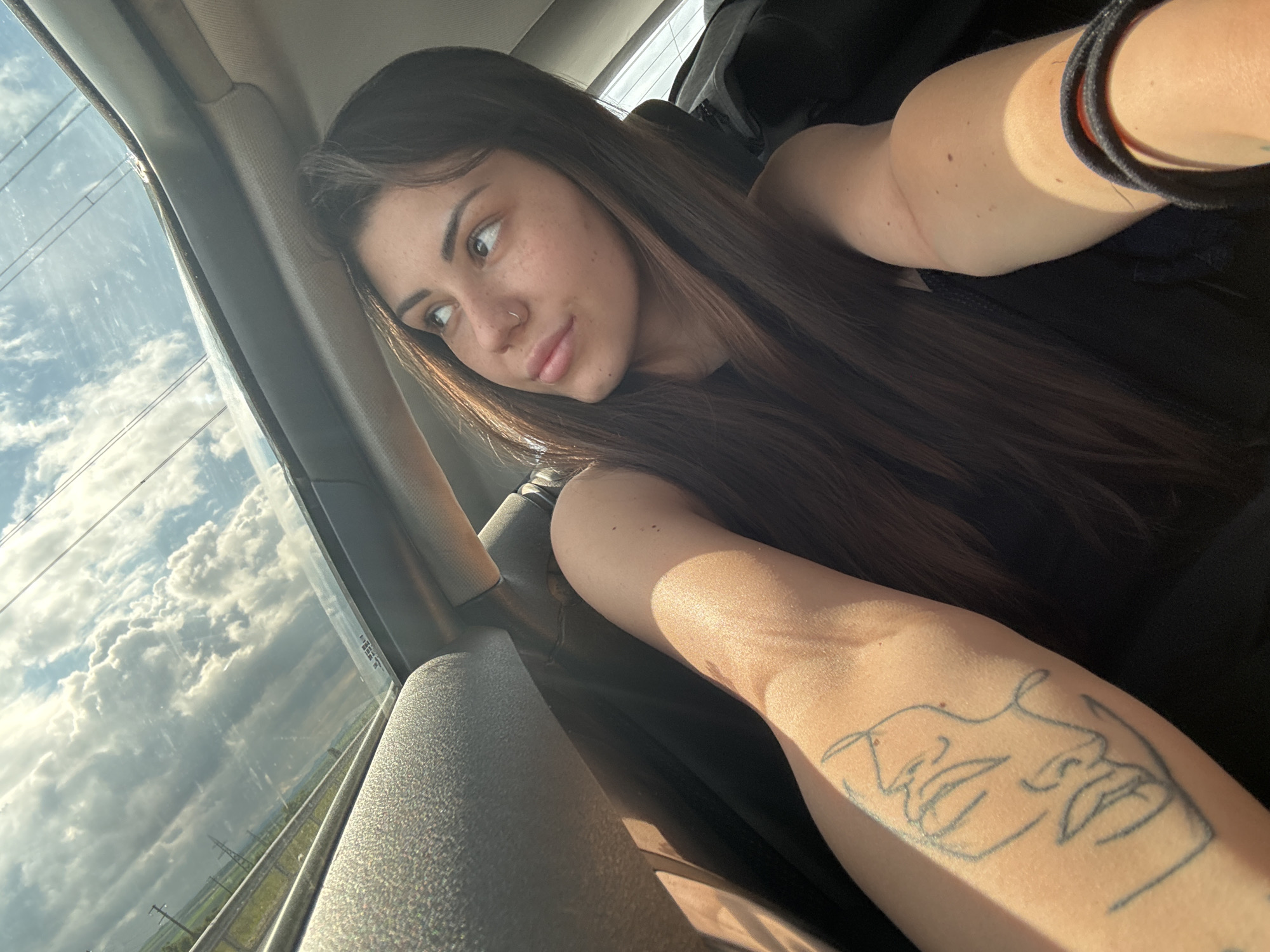 A young woman is depicted in a close-up shot from inside a vehicle, gazing out the window to her left. She has long, dark brown hair, numerous freckles on her face, and a nose piercing. Her expression is soft and contemplative, with a subtle smile, as she observes the external environment. She wears a dark top, possibly black.
Sunlight streams into the car, casting highlights on her skin and the interior. Her right forearm, prominently featured in the foreground, bears a line-art tattoo of two abstract faces. A dark hair tie or bracelet is visible on her left wrist.
Through the car window, a bright, partly cloudy sky dominates, filled with large white and grey clouds against a clear blue sky. Below the clouds, a green, seemingly agricultural landscape unfolds, alongside what appears to be a highway or road with guardrails and power lines, suggesting travel through a rural area. The strong, warm light indicates late afternoon or early evening. The scene is set in or around Hrabarsko, Bulgaria, as suggested by the provided context. The woman appears to be a passenger, passively observing the scenery outside. No visible text is present in the image.