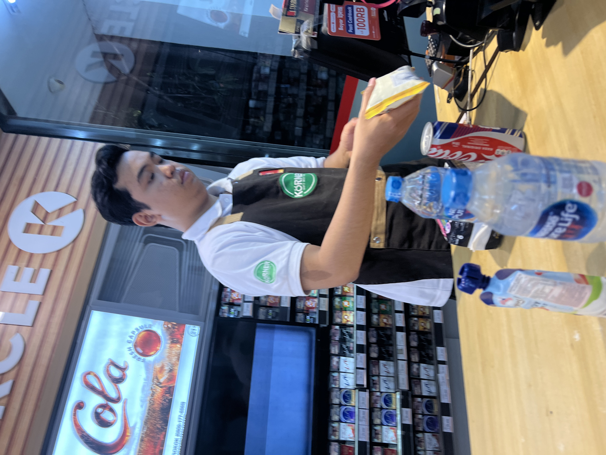 A male cashier stands behind a counter at a Circle K convenience store, preparing to scan items. He wears a white polo shirt with a green "KORNER" logo on the sleeve and a black apron. In his hands, he holds a yellow and white packaged item, likely food or merchandise. On the counter, several items are visible: two bottles of water, one with a blue cap and the other with a white cap and a blue label that reads "Pure Life," and a can of Coca-Cola with red, white, and blue branding. Behind him, shelves are stocked with various cigarette packs. A display above the shelves shows a "Cola" advertisement. The store appears well-lit with overhead lighting. The scene is indoors, typical of a convenience store environment. The location is specified as Denpasar, Indonesia.