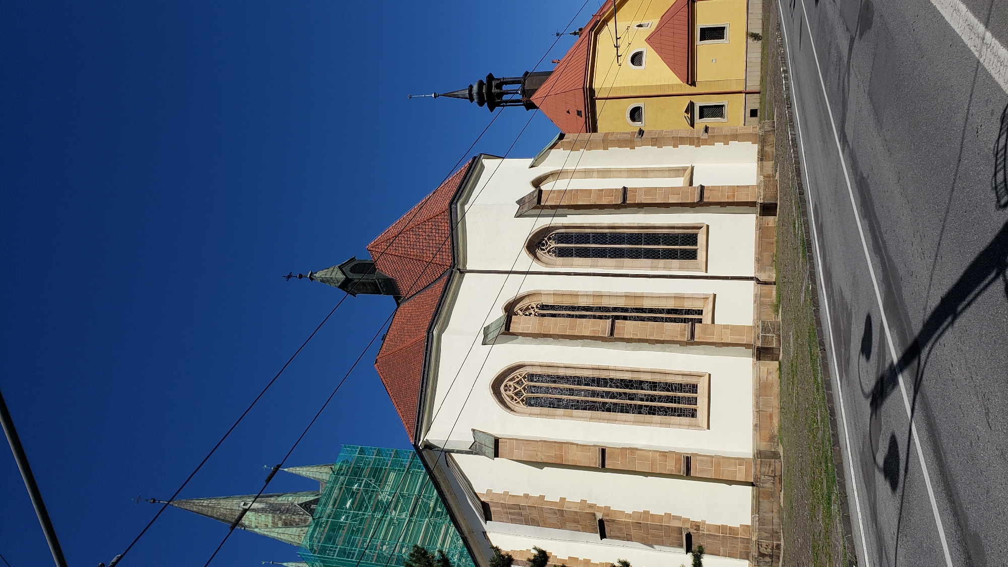 The image depicts a street scene in Prešov, Slovakia, featuring two prominent churches. In the foreground, a light-colored, Gothic-style church with a red tiled roof and tall, arched stained-glass windows dominates the left side of the frame. The church has stone accents along its corners and base. To the right of this church, a smaller, bright yellow building with a red tiled roof and a black spire is visible. In the background, a taller church spire, partially covered in green scaffolding, rises against a clear blue sky. Power lines are strung across the upper portion of the image. The street in the foreground is paved and shows some wet patches, with the shadow of a street lamp cast across it. The environment appears to be an urban setting with the architectural style suggesting a historical district. There are no people or visible animals in the image.