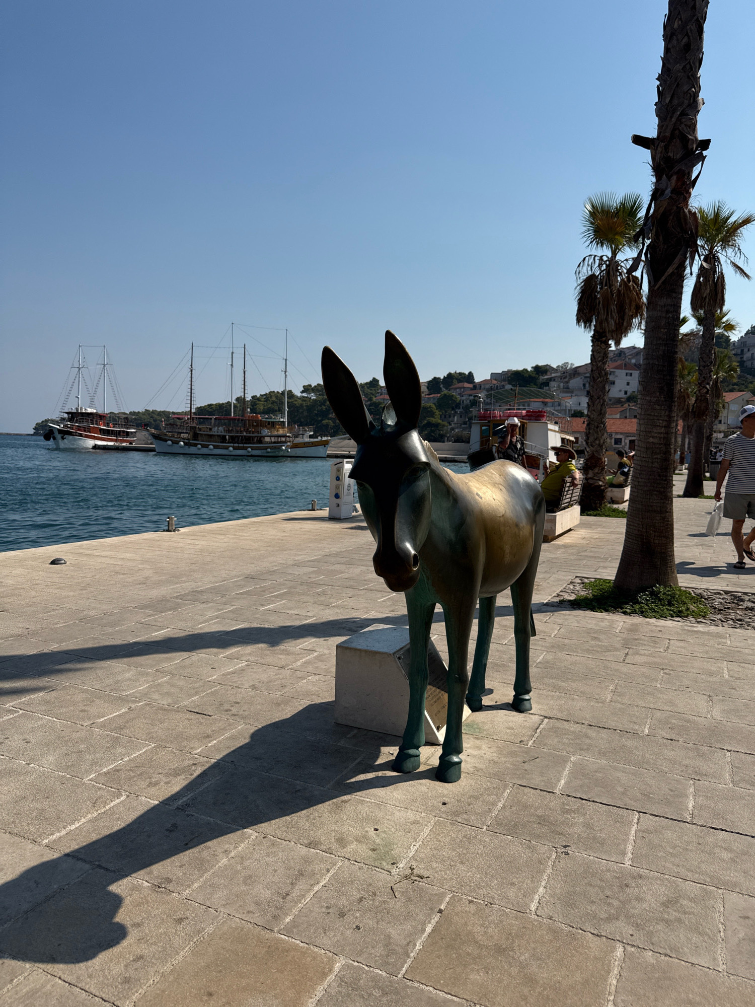 In Jelsa, Croatia, a bronze statue of a donkey stands on a paved walkway near the harbor. The donkey is positioned in the foreground, facing slightly left, with its shadow stretching across the stone tiles. Behind the statue, the calm blue water of the harbor is visible, with several large, multi-masted boats moored. Palm trees line the walkway to the right, and in the distance, white buildings with red-tiled roofs are nestled on a hillside. A few people are seen in the midground, some sitting on benches and others walking, enjoying the sunny day. The sky is clear and blue.