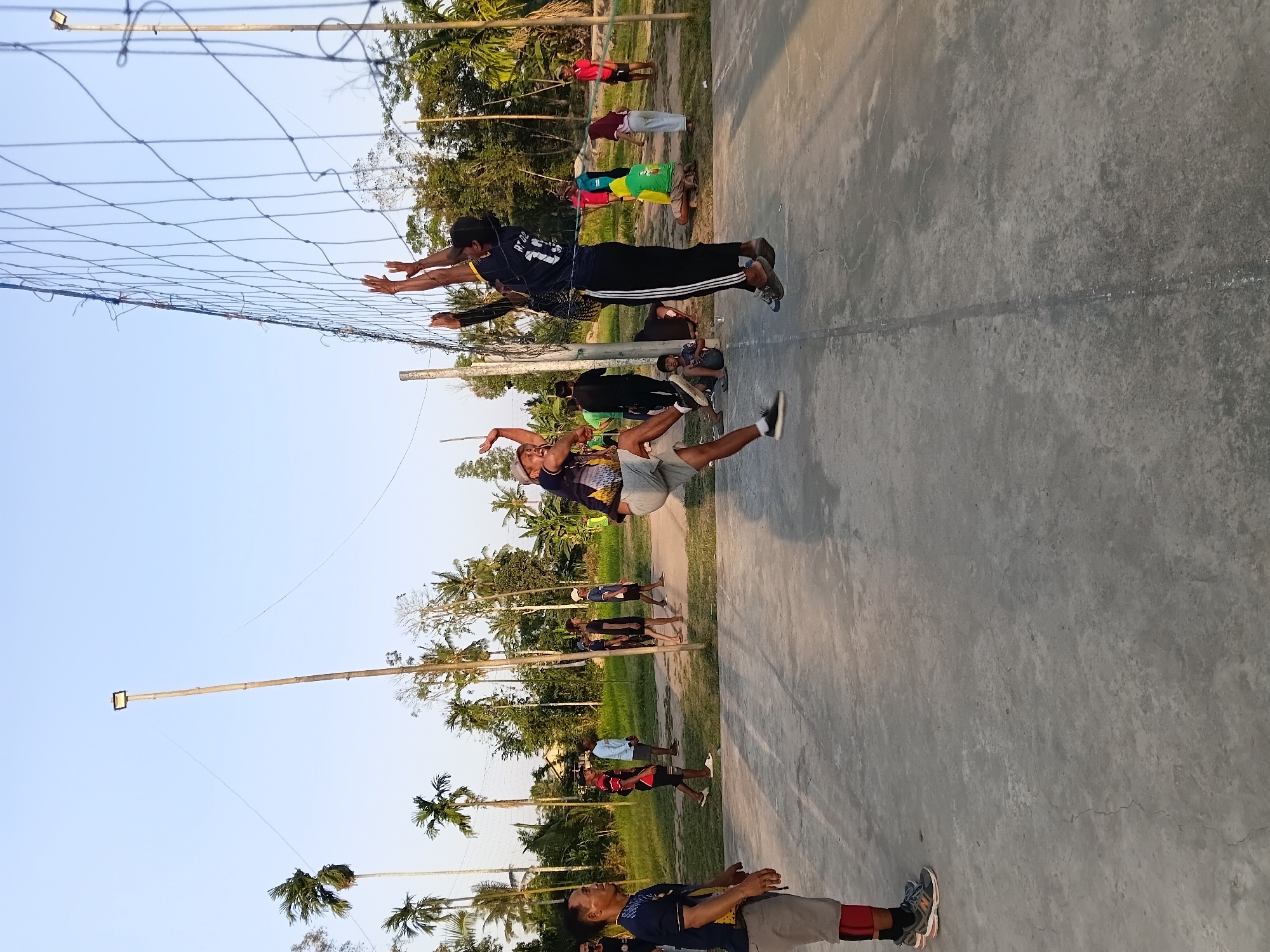 The image depicts a volleyball game taking place on an outdoor court in Margo Rukun, Indonesia. Two men are in mid-air, actively playing volleyball, with one reaching to spike the ball and the other attempting to block. The court appears to be made of concrete. A volleyball net stretches across the frame, dividing the players. Several other individuals are visible on the sidelines, observing the game or in the process of walking around. The environment is characterized by palm trees and other foliage, suggesting a tropical or subtropical setting. A tall pole with a light fixture stands to the left of the frame, and another volleyball net pole is visible in the background. The sky is clear and blue, indicating fair weather.