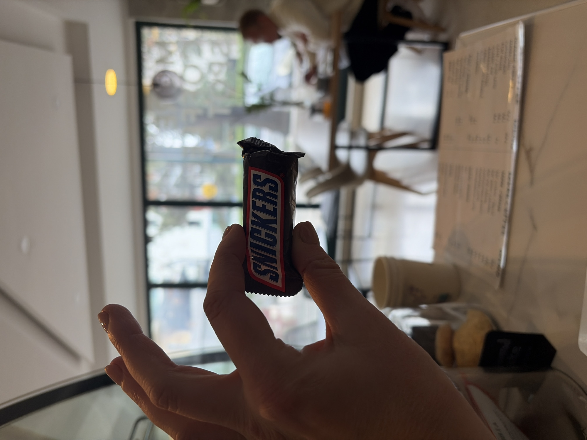 A person is holding a Snickers bar in their fingers, with the word "Snickers" clearly visible on the wrapper. In the background, a person is sitting at a table near a window. The location appears to be inside a cafe or restaurant, as evidenced by the table, menu, and other objects on the table. Considering the context and visual details, the location is the city of Budva, Montenegro.
