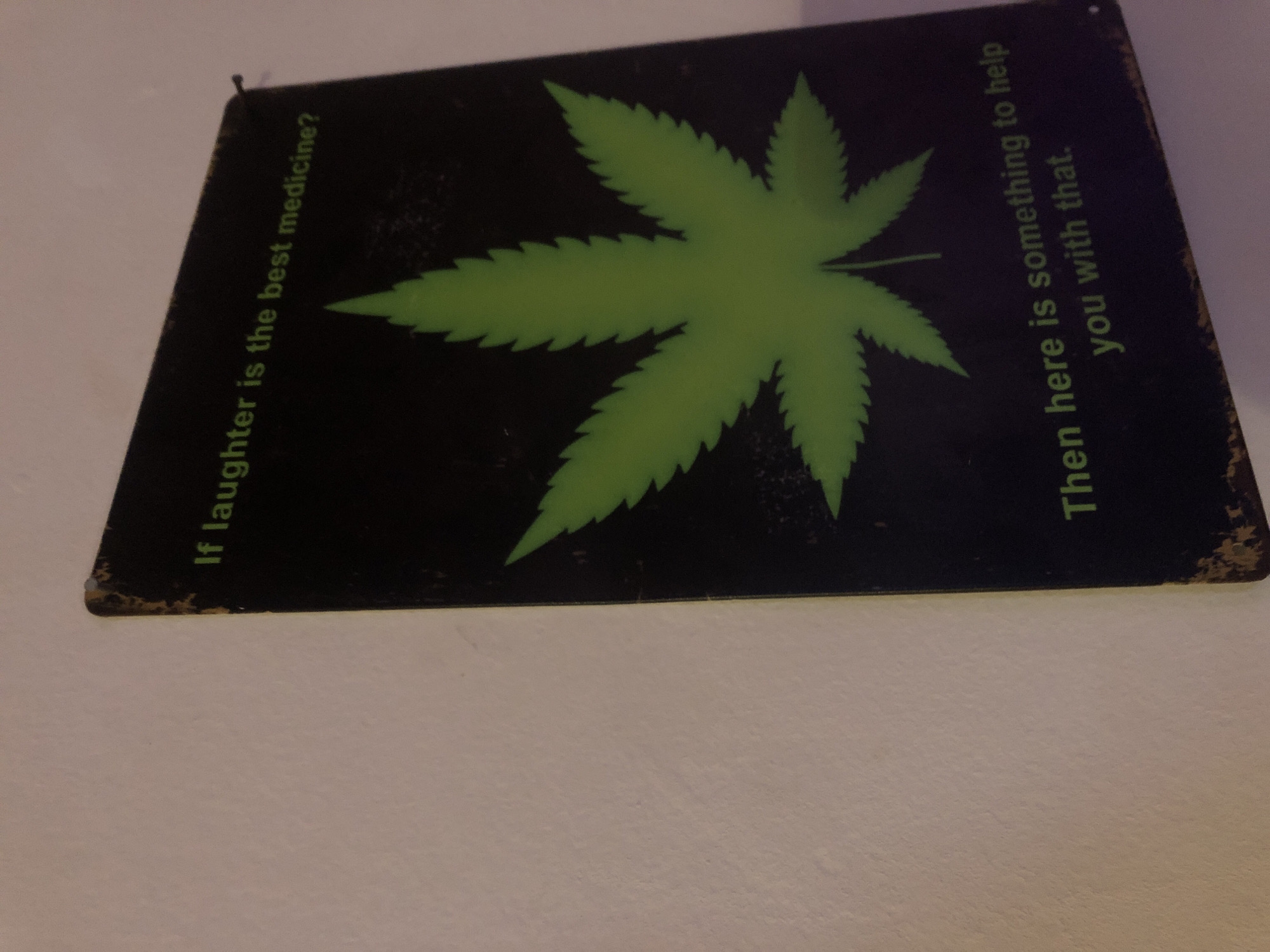 The image shows a black sign with a green cannabis leaf in the center. To the left, it reads "If laughter is the best medicine?" To the right, it reads "Then here is something to help you with that." The sign appears slightly worn around the edges and is attached to a white surface, likely a wall. The image does not contain information about the city of Buenos Aires or Argentina.