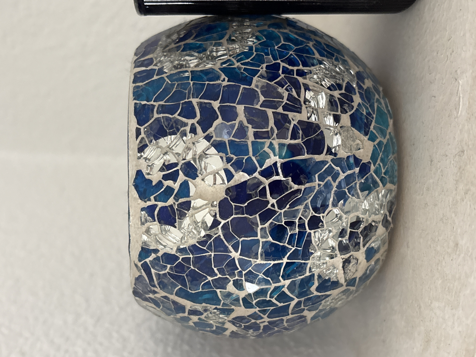 A mosaic glass bowl, predominantly blue and silver, rests on a white surface. The bowl is spherical and has a textured mosaic pattern made of small, irregular pieces of glass. The background is a textured white wall. To the right of the bowl, a portion of a black cylindrical object is visible, likely another object on the shelf. The scene appears to be indoors, possibly on a shelf or table, given the lighting and the white textured background that resembles a wall. There is no indication of the city of Orihuela, Spain, or any people.