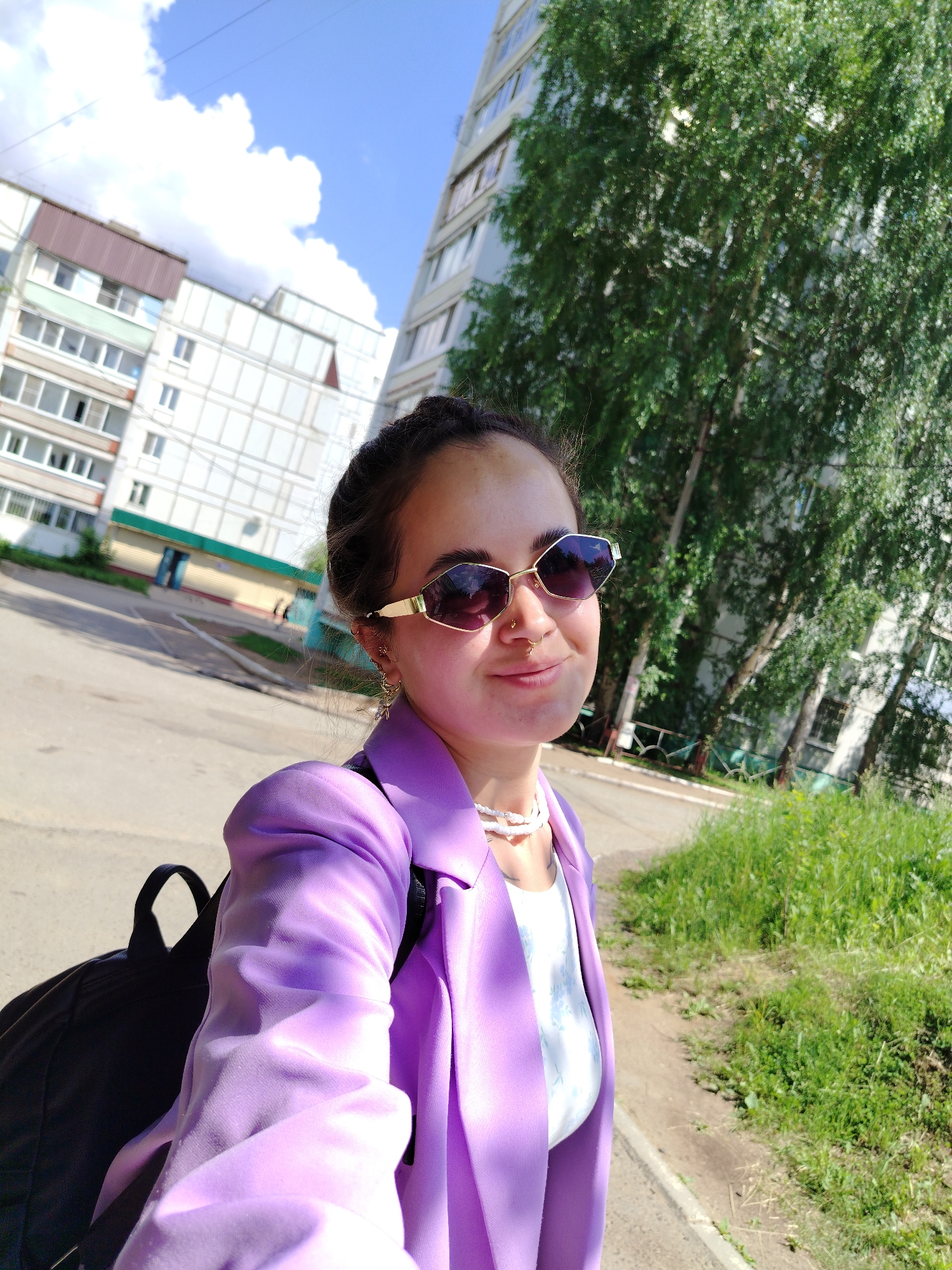 A young woman with dark hair pulled back in a braid is taking a selfie outdoors in Chaikovsky, Russia. She is wearing a purple blazer, sunglasses, a white top with a floral pattern, and a multi-strand white beaded necklace. A black backpack is visible over her left shoulder. The background features a tall, light-colored apartment building and a mature tree with green foliage. The ground is paved, and there is a strip of green grass on the right side of the image. The sky is blue with scattered white clouds.