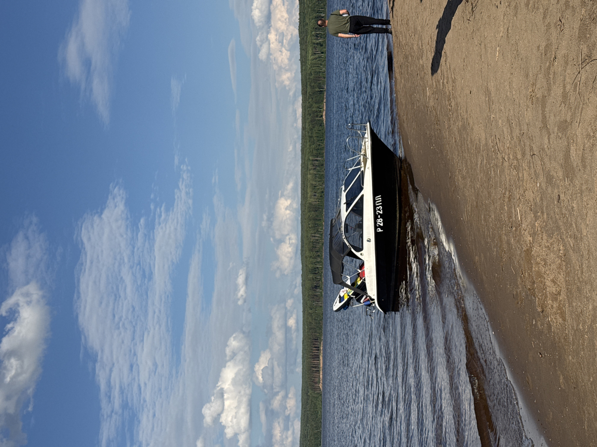 The media file depicts a scenic view of a boat docked near a sandy shore on a body of water. A person, dressed in a green t-shirt and black pants, stands on the shore, facing away from the camera and towards the water. The boat, a modern vessel with a black hull and white cabin, is partially submerged in the shallow water, its bow pointing towards the shore. The water is a deep blue, reflecting the bright sky. The shore is sandy, with some dry vegetation scattered on it. In the background, a dense forest stretches along the horizon, with trees appearing as a dark green band against the sky. The sky is a clear blue with scattered white clouds, suggesting a pleasant day. The overall scene conveys a sense of tranquility and outdoor leisure. The time of day appears to be daytime, likely late morning or early afternoon, given the brightness of the sun and the shadows cast. The location cues, such as the type of vegetation and the overall landscape, are consistent with a natural environment like a lake or river in a temperate region. Visible text on the boat reads "Р 28-23 ПЛ".