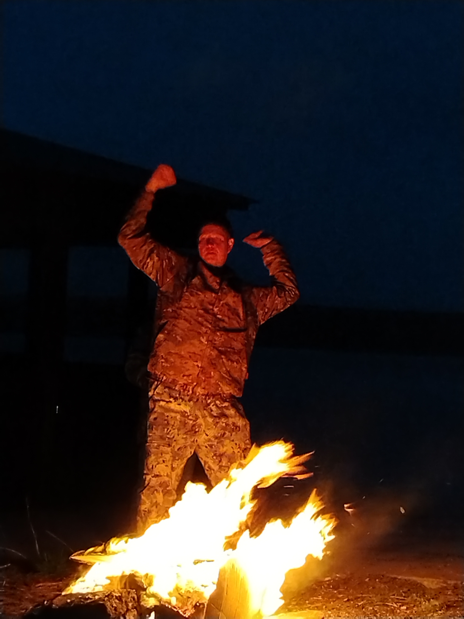 The image captures a man in a camouflage uniform standing next to a large, bright bonfire at night in an outdoor setting in Chaikovsky, Russia. The man is positioned with his arms raised and bent at the elbows, hands seemingly clenched, in a dynamic and energetic pose, looking towards the viewer. His face and the front of his uniform are intensely illuminated by the warm, orange-red glow emanating from the roaring flames below.
The bonfire, dominating the lower part of the frame, consists of burning logs or debris, casting strong light and shadows across the scene. The background is a very dark, indistinct blue-black, indicative of deep night or late twilight, with faint, unidentifiable dark structures or natural elements visible in the far distance. The ground around the fire appears to be dark earth with some scattered debris. The overall mood conveyed by the pose and the dramatic lighting from the fire suggests excitement, triumph, or celebration. No text is visible in the image.