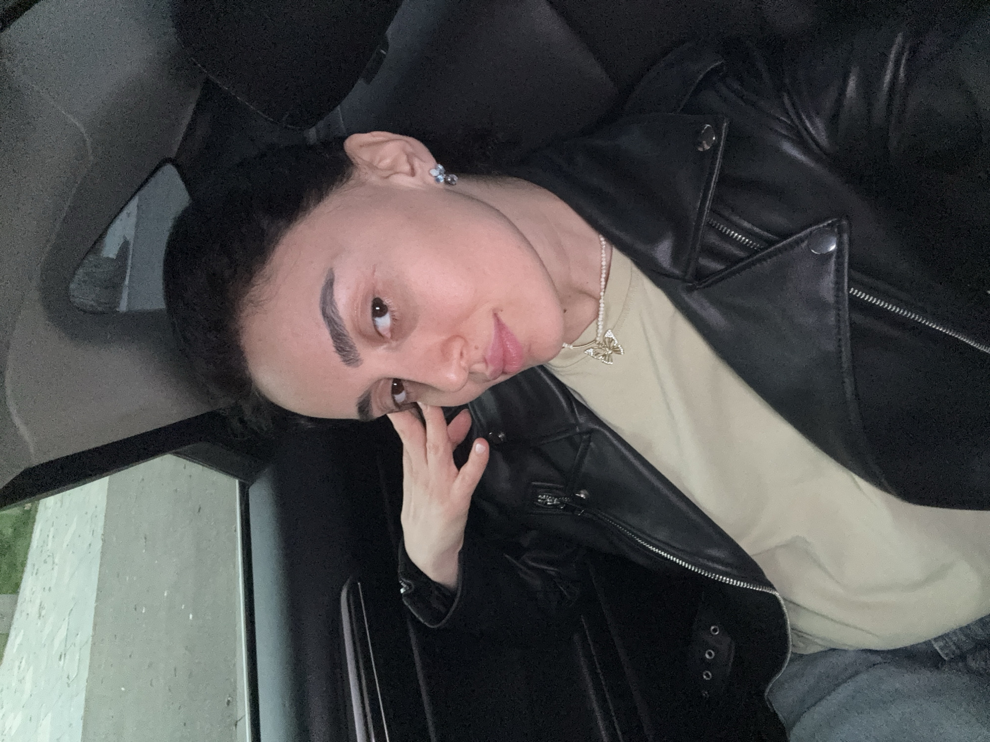 A woman is depicted from a medium close-up perspective, seated in the rear passenger compartment of a vehicle. Her dark hair is pulled back, revealing her face. She possesses prominent eyebrows, light brown eyes, and her lips are slightly parted. She is wearing a black leather jacket over a light olive-beige t-shirt, a small pearl choker necklace adorned with a gold-colored butterfly pendant, and light-toned stud earrings. Her left hand is raised, with fingers gently touching the side of her forehead near her temple. She maintains direct eye contact with the camera. The vehicle's interior features dark upholstery and dark paneling on the door to the left. Through the partially visible window on the left, a blurred outdoor environment is discernible, characterized by a gray, possibly paved, surface and indistinct green areas, suggesting an outdoor setting in ambient light. The precise location within Tbilisi, Georgia, cannot be determined from the visible external environment.