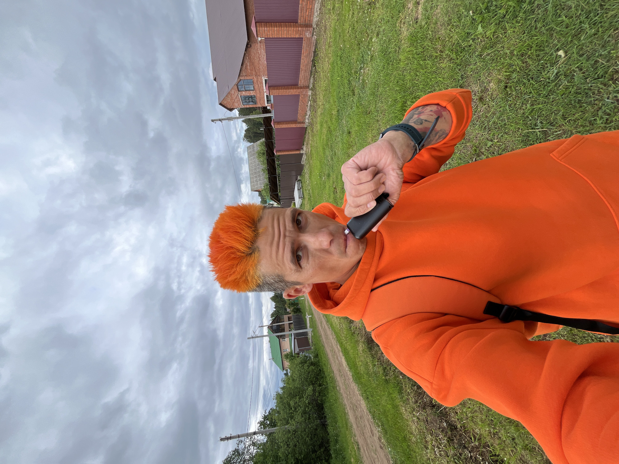 A person with distinctive bright orange, spiky hair is featured in a close-up, selfie-style photograph taken outdoors. They are wearing a vibrant orange hooded sweatshirt, and their left forearm, visible from the sleeve, displays tattoos and a dark-colored wristband or watch. The individual holds a black vape pen or e-cigarette to their mouth, suggesting they are either vaping or about to do so. Their expression is direct, looking at the camera with a neutral to slightly serious gaze.
The scene is set in a rural or semi-rural area under an overcast sky filled with gray clouds, indicating a cloudy daytime. A narrow dirt road stretches through a grassy expanse in the background, bordered by green trees and bushes. Several buildings are visible, including a brick house with a brown roof and a brown fence or gate, and another house with a green roof located further down the road. Utility poles and power lines are also present, adding to the landscape details typical of the provided location context of Chaikovsky, Russia. No visible text is present in the image.