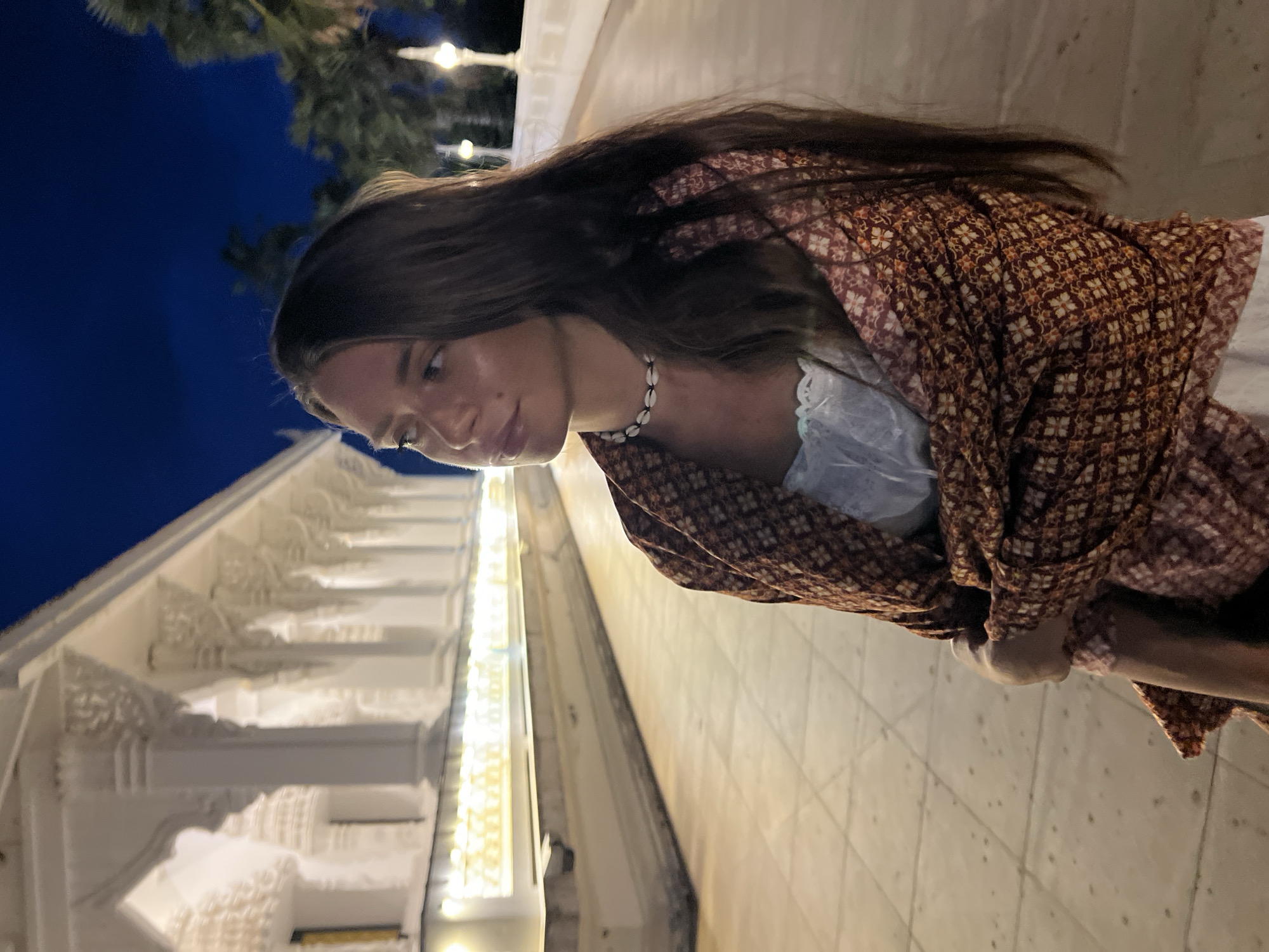 A woman with long brown hair and wearing a patterned orange and brown kimono-style jacket and a seashell necklace stands outdoors, likely in Baan Bo Phut, Thailand. She is looking down. To her left is a white, ornate building illuminated with lights. The background sky is dark blue. The surface on which she is standing appears to be tiled.