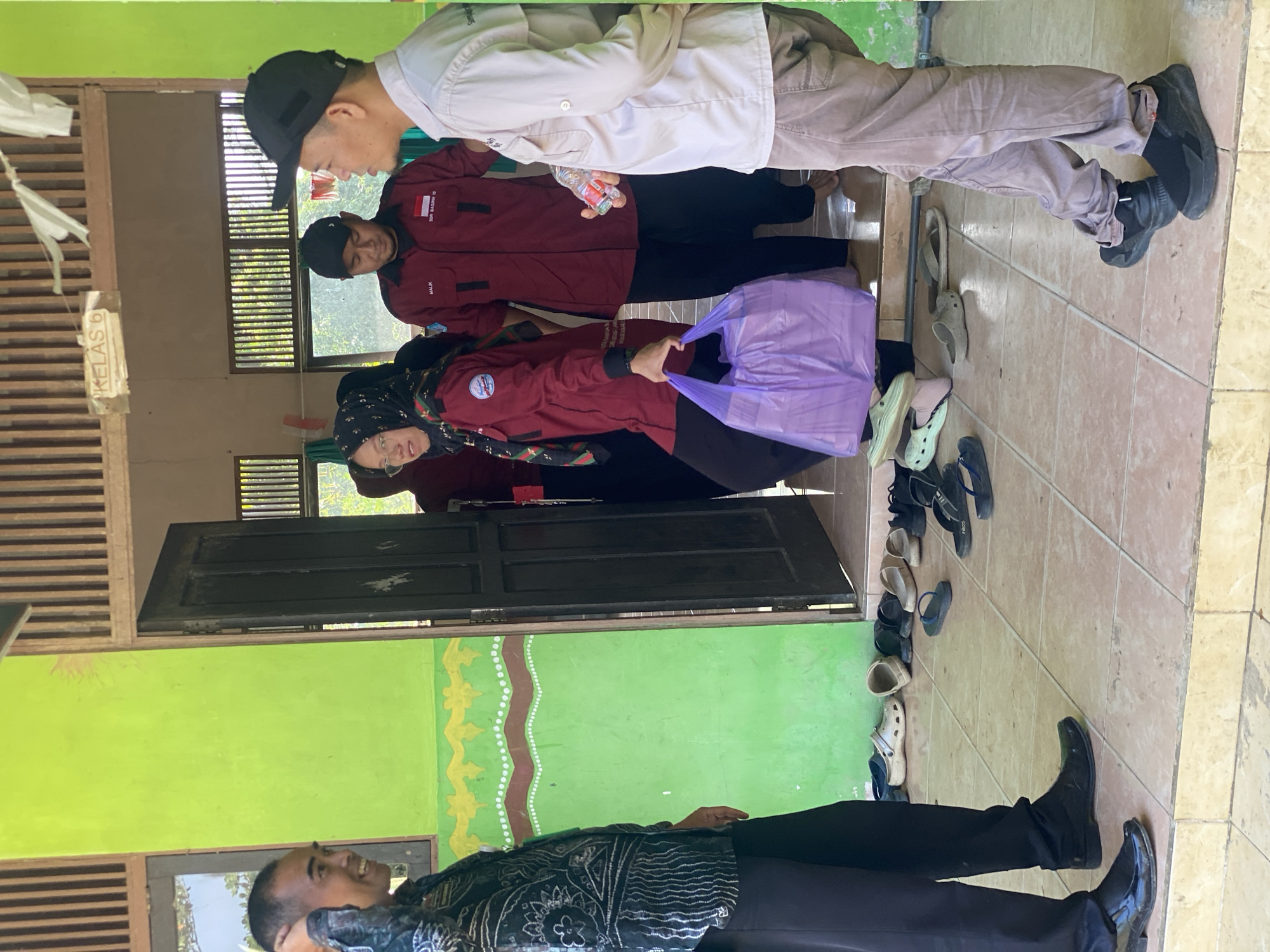 In Handil Baru, Indonesia, a group of individuals stands on a tiled porch outside a building with a lime green exterior and a wooden doorframe. A sign above the doorway reads "KELAS 6," indicating it is a classroom. Four people are visible. On the left, a man in a dark patterned shirt and black pants smiles and touches his head, facing two people entering the doorway. Inside the doorway, a woman in a maroon jacket, headscarf, and glasses holds a large purple plastic bag filled with items. To her right, a man in a maroon jacket with the Indonesian flag and text on it, and another man in a light-colored shirt and brown pants are visible. The man in the maroon jacket is looking down, and the man in the light-colored shirt is speaking to the man on the left. Various shoes, including sandals and crocs, are neatly arranged on the floor near the entrance, suggesting a practice of removing footwear before entering. The environment appears to be outdoors, with sunlight casting shadows on the tiled floor.