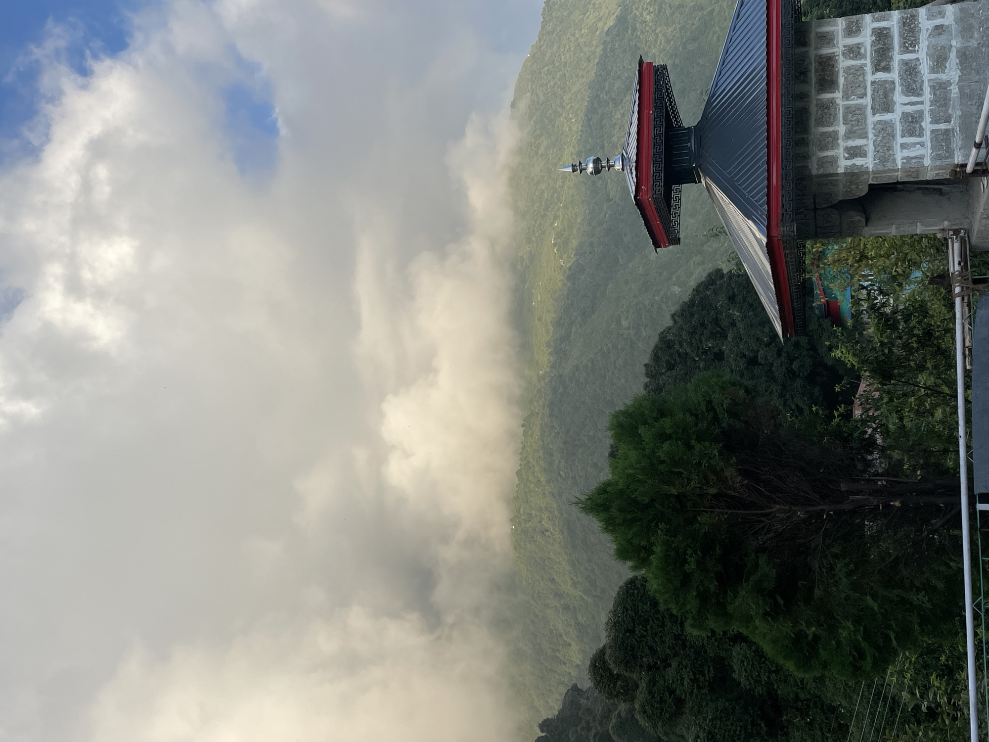 The image depicts a mountainous landscape with a Tibetan-style structure in the foreground. The structure is made of grey stone blocks and features a dark-colored, multi-tiered roof with red trim. A pointed, metallic finial adorns the highest point of the roof. Behind the structure, lush green hills are visible, partially obscured by thick, white clouds that are illuminated by sunlight. Dense green trees populate the lower slopes of the hills and the area around the structure. The sky above is a mixture of white clouds and patches of blue. The scene appears to be an outdoor setting in a hilly region. The style of architecture and the surrounding vegetation suggest a location in the Himalayan foothills, possibly in Bhagsu, India, known for its Tibetan culture and monasteries. There are no people visible in the image.