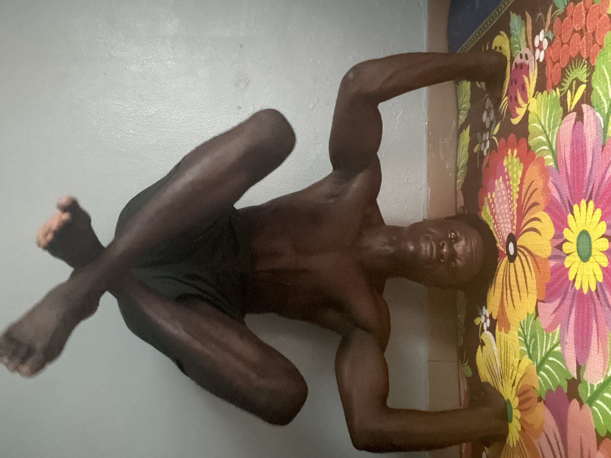 A man with dark skin and muscular physique is lying on his back, with his legs crossed. He is wearing dark shorts. He is positioned on a light-colored floor, next to a colorful rug with a floral pattern. The rug features large, vibrant flowers in shades of pink, yellow, orange, and red, with green leaves. The man's arms are extended, one reaching towards the rug and the other resting behind his head. His head is turned towards the camera, and he appears to be looking slightly upwards. The background consists of a plain, light-colored wall. The lighting suggests it might be daytime. The scene appears to be indoors, possibly in a home in Badagry, Nigeria. There is no visible text in the image.