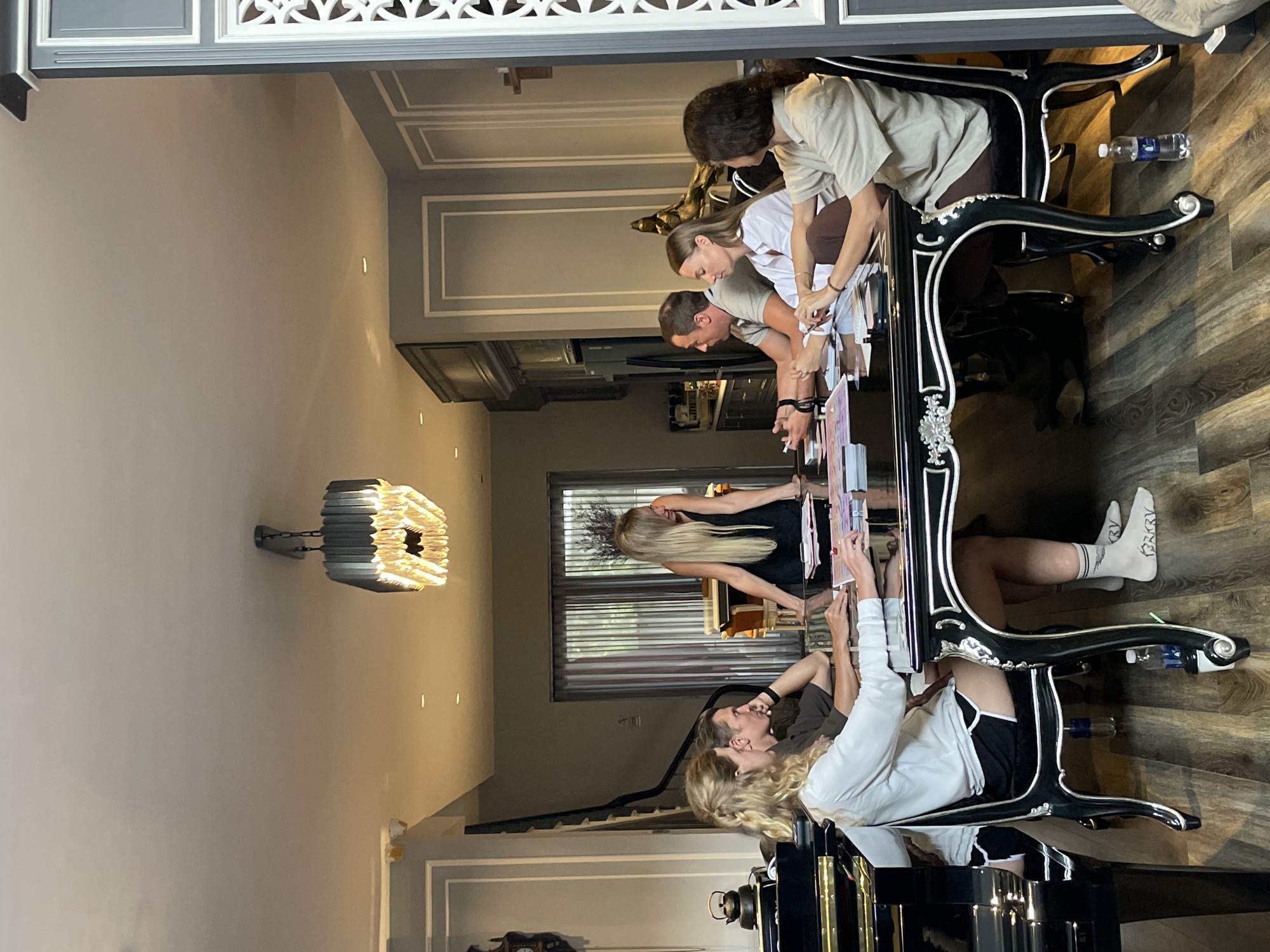 The image depicts a group of young adults gathered around a large, ornate black table, likely a dining or conference table, in what appears to be a well-decorated indoor setting. The room has light-colored walls with elegant molding, and a distinctive, modern square chandelier hangs from the ceiling. The floor is made of dark wood planks.
There are at least six individuals visible, some of whom are intently focused on papers or objects spread across the table, suggesting an activity like a meeting, study session, or collaborative project. One person is wearing white socks with black lettering that appears to say "GRY". Another individual has their arm resting on the table, holding a pen. A person with long blonde hair is leaning forward, examining something on the table.
The lighting suggests it could be daytime, with natural light possibly coming from a window visible in the background, which has blinds. The overall atmosphere seems engaged and focused. The image is taken from a slightly elevated, rotated perspective, giving a dynamic view of the scene.