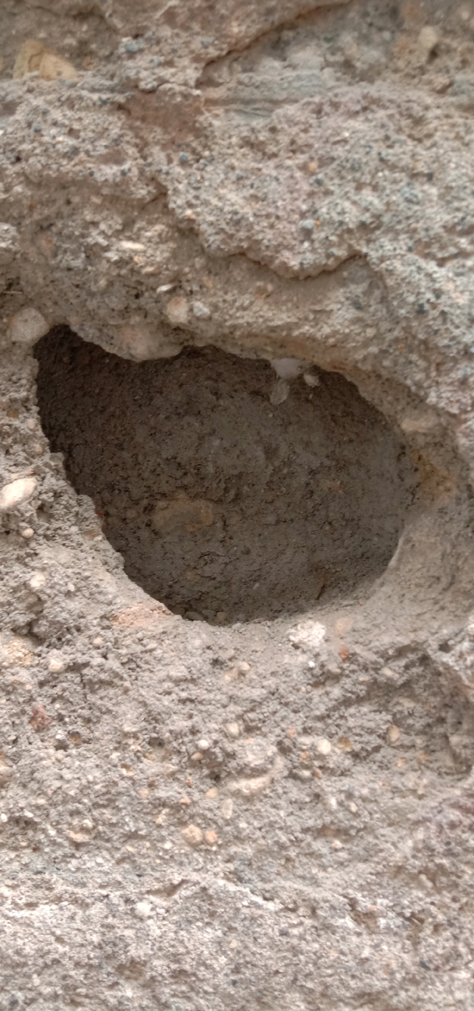 The image shows a close-up, eye-level, full shot of a damaged concrete surface with a hole. The texture of the concrete is rough and granular, with visible aggregate pieces. The hole is irregular in shape and appears to be deep, with its interior also composed of concrete. The lighting is consistent, suggesting an outdoor environment, but no specific location in Tangerang, Indonesia, can be identified from the image alone. There are no people, animals, or any other identifiable objects or actions present. The focus is solely on the textural details and the void within the concrete structure.