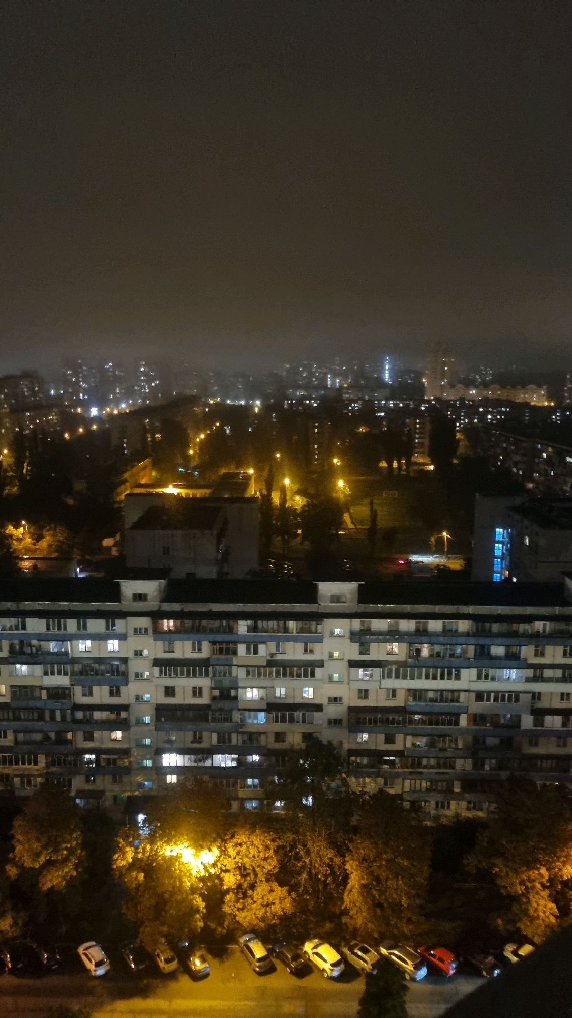 This is a nighttime aerial view of a residential building in Kyiv, Ukraine, likely a Soviet-era apartment block. The building is large and imposing, with numerous windows, many of which are lit, indicating occupancy. In front of the building, there is a row of parked cars, some illuminated by streetlights. The background shows a cityscape with more buildings and lights, suggesting an urban environment. Trees are also visible, their leaves illuminated by artificial light, creating a warm glow. The sky is dark and overcast, contributing to the moody, nighttime atmosphere.