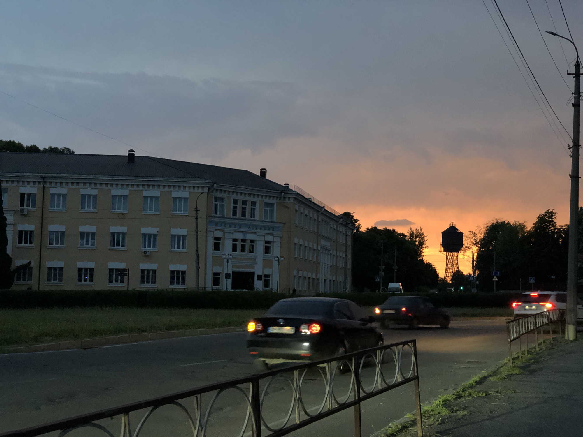 The image depicts a street scene at dusk in an urban setting. A large, multi-story building with a light yellow facade and numerous windows is visible on the left side of the frame. Cars are driving on a road in front of the building, with some showing motion blur, indicating they are in motion. The sky is a dramatic blend of dark gray clouds and bright orange hues, characteristic of a sunset or sunrise. In the distance, a tall water tower with a dark cylindrical tank and a lattice-like support structure stands against the colorful sky, silhouetted by the light. Trees line the horizon behind the road and the building. A street lamp on a utility pole is present on the right side of the image, with power lines extending across the frame. A metal railing with a circular design runs along the bottom foreground of the image, separating the sidewalk or pedestrian area from the road. The overall atmosphere is one of a city winding down for the evening, with the natural beauty of the sky providing a striking contrast to the urban environment.