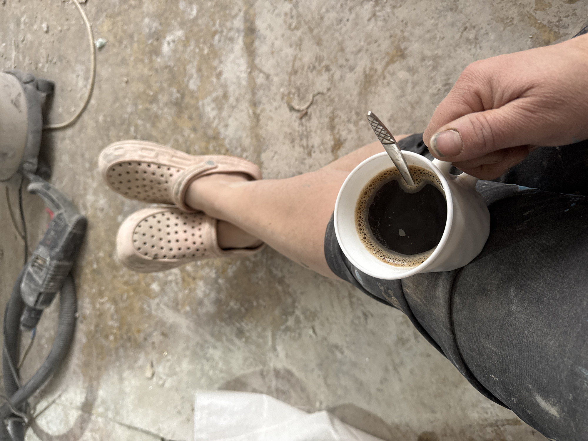 The image shows a person sitting with legs crossed, wearing pink perforated clogs. A white mug filled with dark coffee is held in their right hand, a small spoon resting inside. The person is wearing dark-colored pants that are smudged with what appears to be white dust. To the left, a gray vacuum cleaner or similar power tool lies on the dusty, light-colored floor. The location is potentially a construction or renovation site in the city of Chaikovsky, Russia.