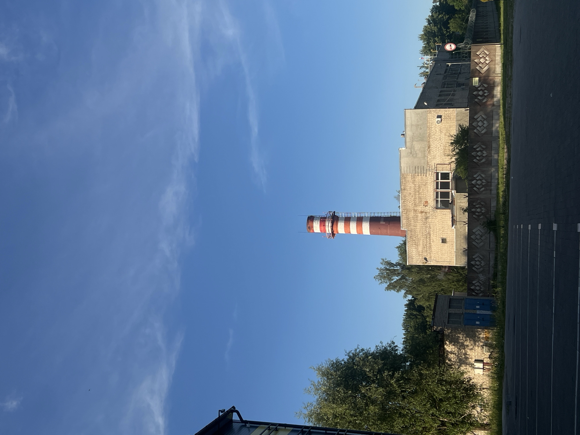 The image shows an industrial area with a tall, red and white striped smokestack dominating the center-left of the frame. To the right of the smokestack is a light-colored industrial building with several windows. In front of the building is a grey concrete fence with a decorative diamond pattern. To the left of the smokestack, a large green tree obscures part of another smaller building, which appears to be constructed from brick and has blue doors. The foreground is a paved area with white lines, suggesting a parking lot or road. The background consists of a dense line of trees against a clear blue sky with wispy white clouds. A small bird is visible in the sky. A circular sign with a red border and two red horizontal bars is present on the right side of the image, indicating a prohibition. The overall scene is an outdoor industrial setting bathed in sunlight, likely in the city of Kaliningrad, Russia.