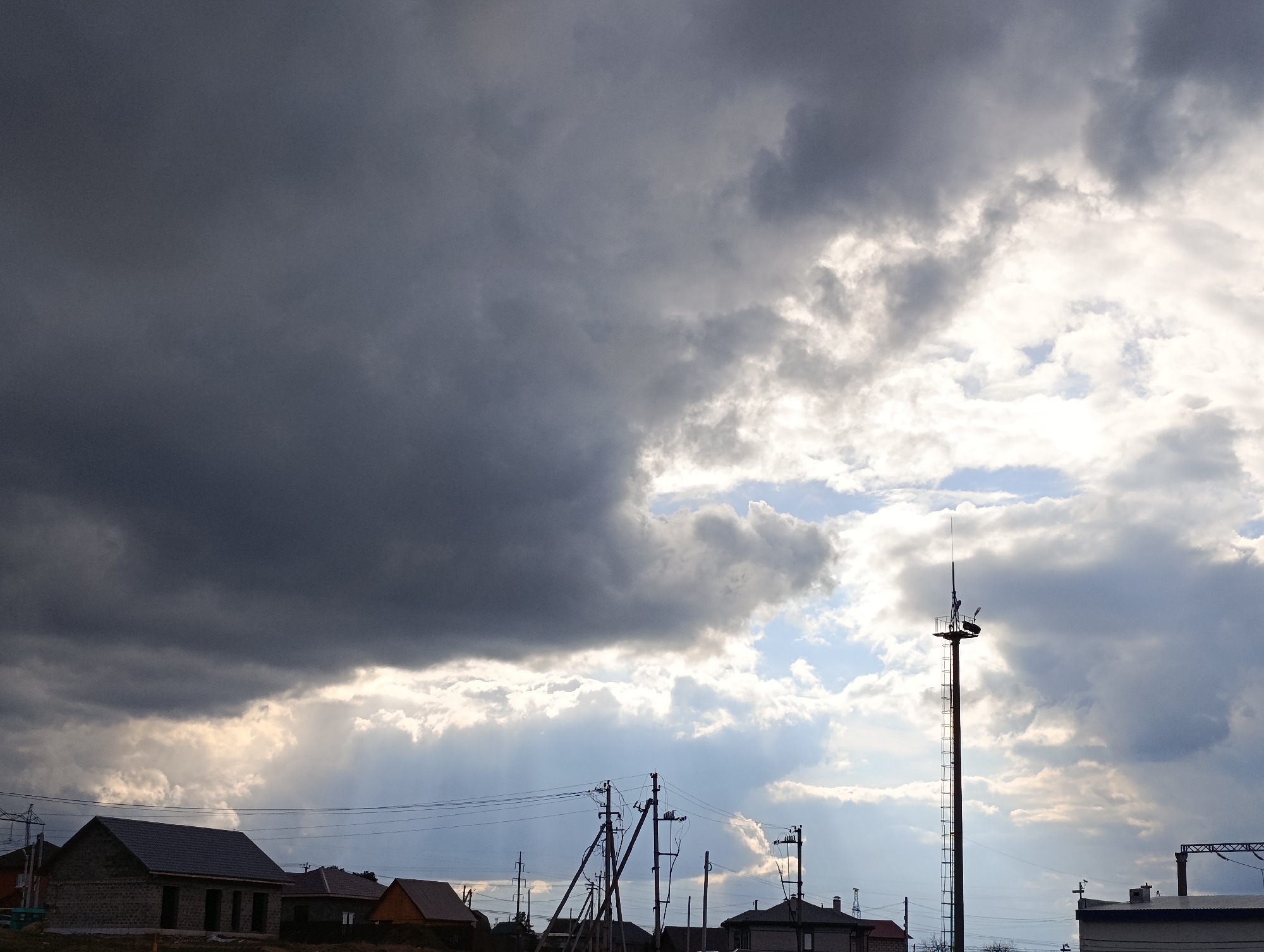 The image depicts a cloudy sky over a residential area. The sky is dominated by dark, ominous clouds on the left side, transitioning to lighter, brighter clouds on the right, where glimpses of blue sky are visible. A tall metal tower stands prominently in the center-right of the frame, with a platform and a ladder extending up its length. Power lines stretch across the scene in the middle ground, connecting to utility poles. Several buildings are visible at ground level, including a brick building under construction, houses with varying roof colors, and a larger, partially obscured building on the right edge. The scene appears to be in Chaikovsky, Russia.