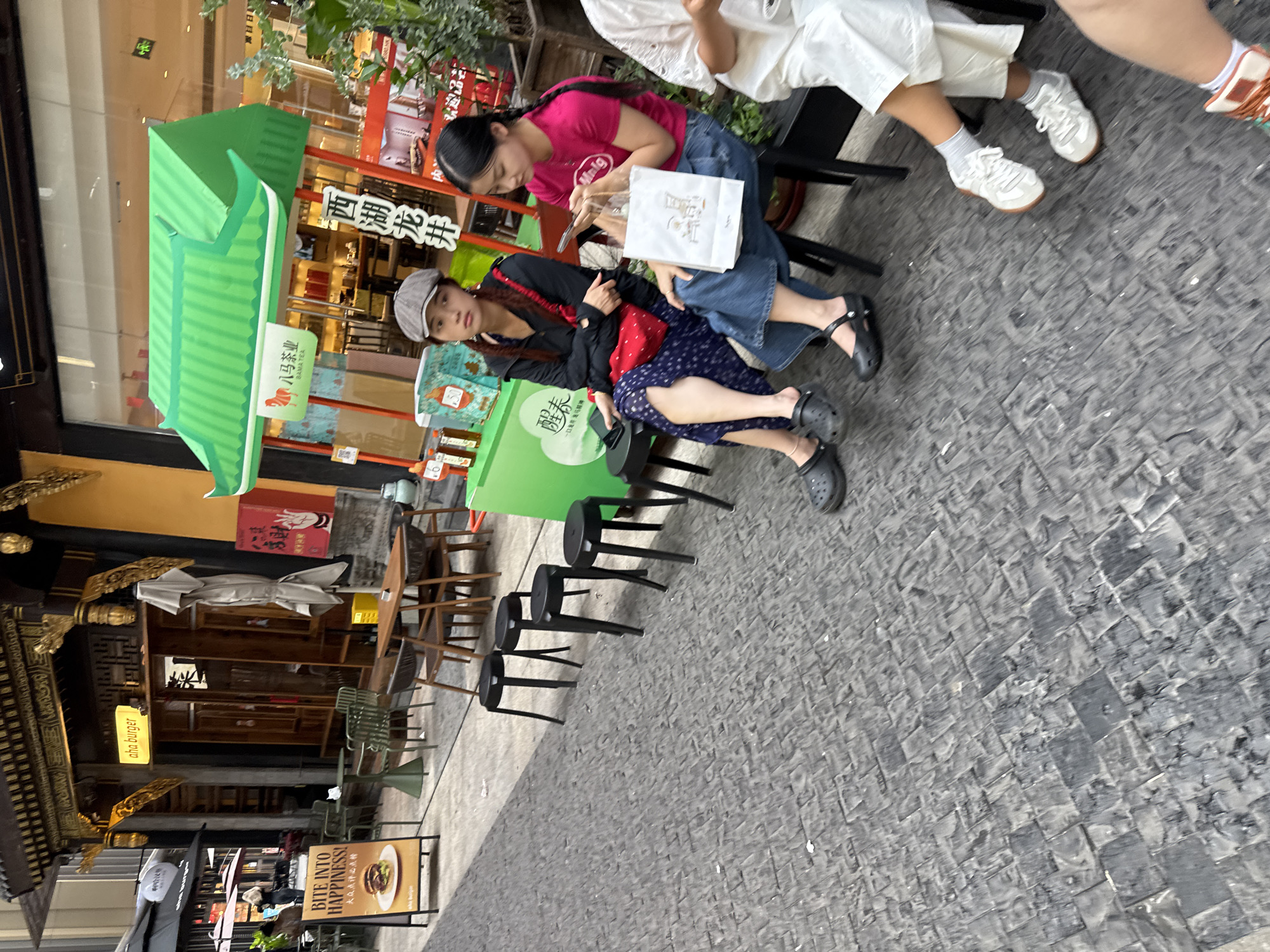 The image depicts an outdoor scene on a paved street in Yuzhong District, China. Two young women are seated on black stools in the foreground. The woman on the left, wearing a grey cap, black jacket, and a blue and white polka dot skirt, has a red bag on her lap. The woman on the right, wearing a bright pink t-shirt and blue jeans, is looking at her phone and holding a white paper bag. To the left of the women is a green food stall with Chinese characters and a red sign. Behind the stall is a building with traditional Chinese architectural elements, including a gold-trimmed roof and wooden doors. A sign for "aha burger" is visible above the entrance of this building. In the background, several tables and chairs are set up outside, suggesting a dining area. The ground is paved with dark, textured tiles.