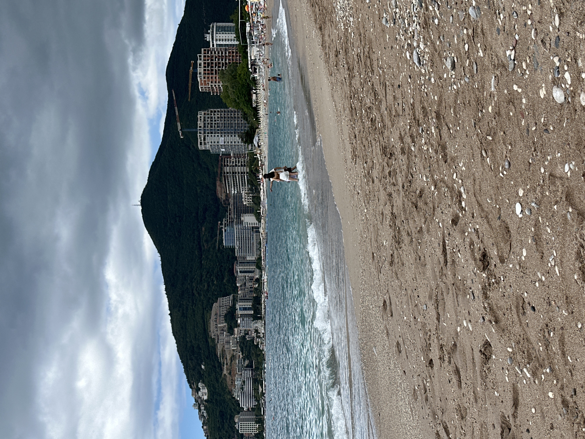 This media file captures a panoramic view of a coastal scene in Budva, Montenegro. The image showcases a sandy beach, the sea, a prominent mountain range, and extensive urban development under a partially cloudy sky.
In the foreground, a wide stretch of coarse sand is visible, densely covered with small pebbles and shells. Several footprints mark the sand closer to the water. The light blue-green ocean water laps at the shore with gentle waves. Two individuals, appearing to be a couple or friends, are standing in the shallow water facing the sea. Further down the beach, other people can be seen relaxing on sun loungers or swimming.
The midground features a long strip of modern buildings, including what appear to be hotels or apartment complexes, situated along the coastline at the foot of a large mountain. Several structures show signs of ongoing construction, with visible concrete frameworks and construction cranes.
The background is dominated by a steep, dark green, tree-covered mountain that rises dramatically behind the urban development. The sky overhead is a mix of heavy grey clouds and lighter, clearer patches, suggesting variable weather conditions. The scene depicts a lively daytime atmosphere, combining natural beauty with significant human activity and development. No visible text is present in the image.