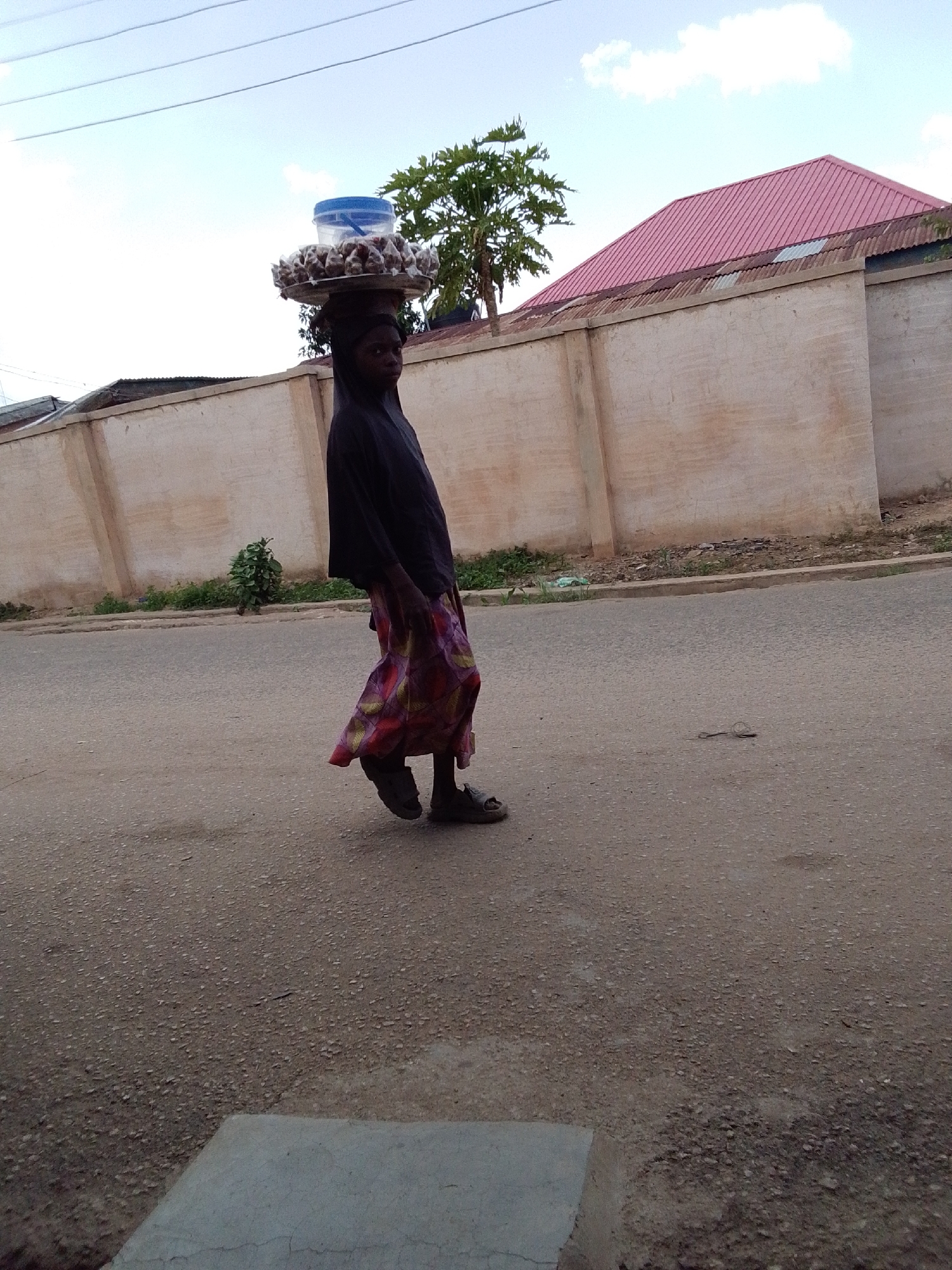 A young person, likely a woman, walks along a paved road in Jos, Nigeria. She carries a large tray balanced on her head, laden with what appears to be food items and topped with a blue plastic container. She is dressed in dark clothing over a patterned skirt. In the background, a tan wall runs along the edge of a property, with a building featuring a red roof visible behind it. A tree stands near the wall. The road is largely empty except for the figure walking.