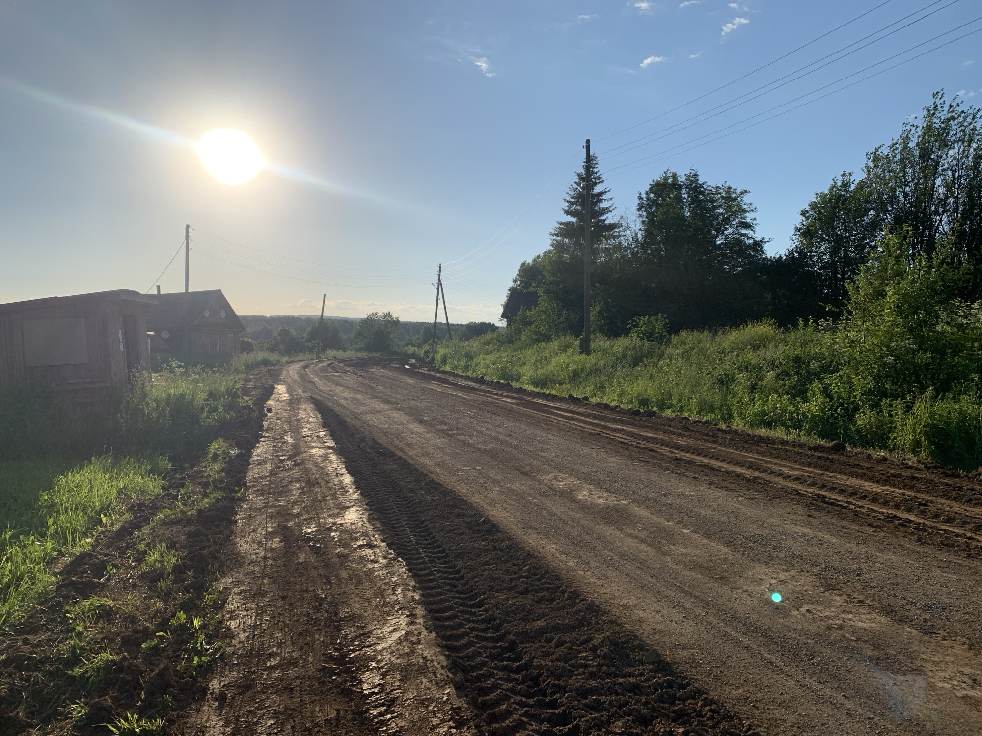 The image depicts a rural dirt road under a bright, sunny sky in Oktyabr'skii, Russia. The sun is prominently featured in the upper left quadrant, casting a strong lens flare across the sky. Power lines stretch across the frame, supported by wooden utility poles. To the left, a weathered wooden building, possibly a house or shed, is visible, surrounded by tall green grass and some disturbed earth. The dirt road itself shows signs of recent activity, with visible tire tracks and areas of mud and loose soil, suggesting construction or maintenance work. The road curves and recedes into the distance, bordered by lush green vegetation and trees on the right side. The overall environment is that of a country setting during daylight hours.
