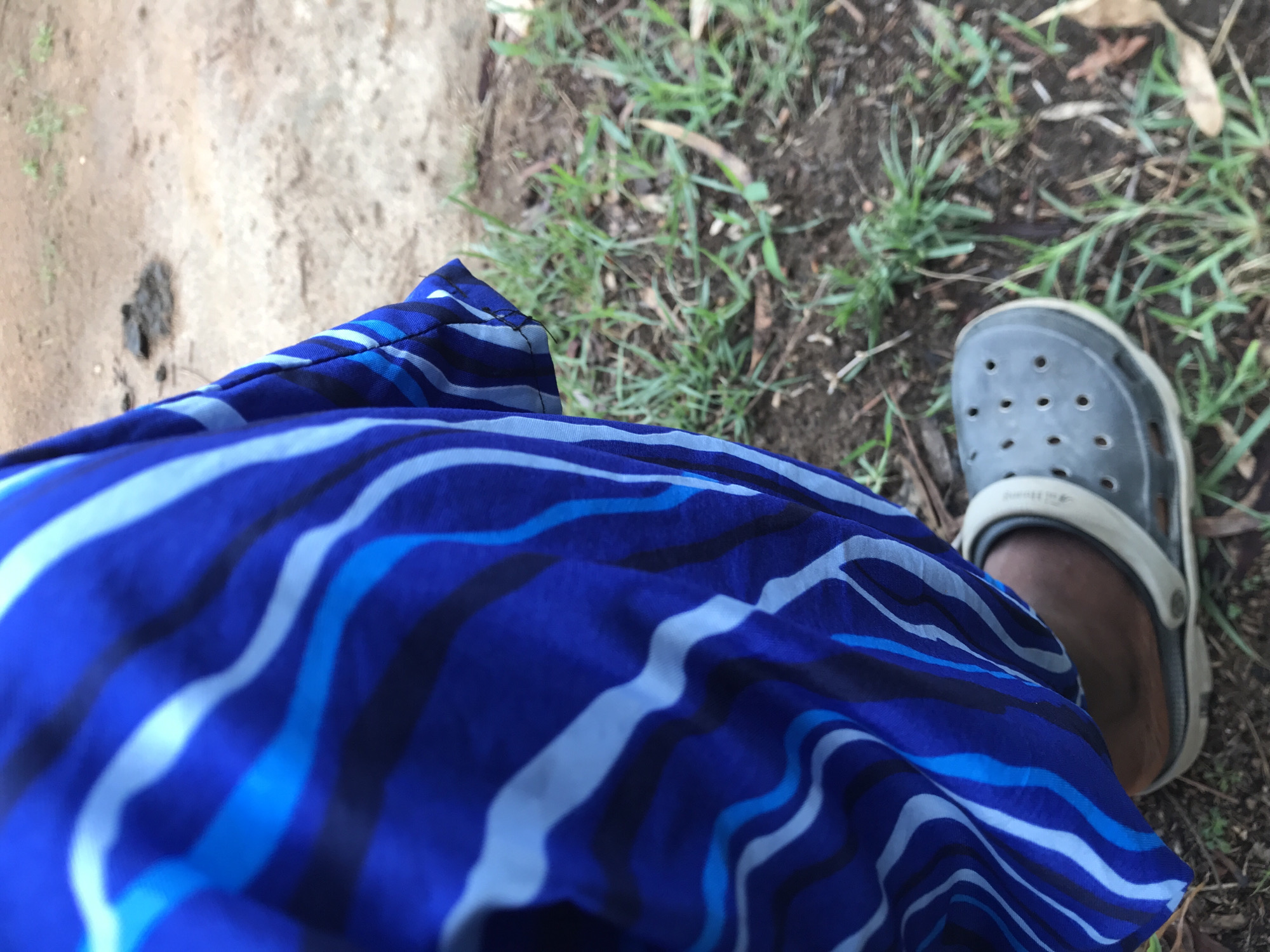 The image presents a high-angle, downward-looking perspective of a person's lower body in an outdoor setting. The central focus is a vibrant blue garment, possibly a skirt or wrapper, adorned with a dynamic pattern of wavy stripes in various shades of blue, light blue, and white, with some darker lines.
The person's right foot, featuring dark skin, is visible wearing a grey Croc-style sandal. The sandal has the typical perforated design on top and a lighter-colored, almost cream-white, adjustable strap. Faintly embossed on this strap, the word "CROCS" is discernible.
The ground beneath is a blend of textures and colors. To the left of the garment, there's an expanse of light brown, sandy or dusty earth, with a dark, irregular clump, possibly dried mud or animal droppings. To the right and around the foot, the ground is dark, exposed soil interspersed with patches of green grass and a few fallen, dry leaves. This environment suggests an unpaved area, typical of rural or semi-urban pathways.
The scene depicts a casual moment outdoors, with the person seemingly standing or casually walking. The lighting indicates it is daytime, with soft natural light illuminating the area. The overall context, particularly the unpaved ground and vegetation, aligns with the provided location context of Unguwar Saya, Nigeria.